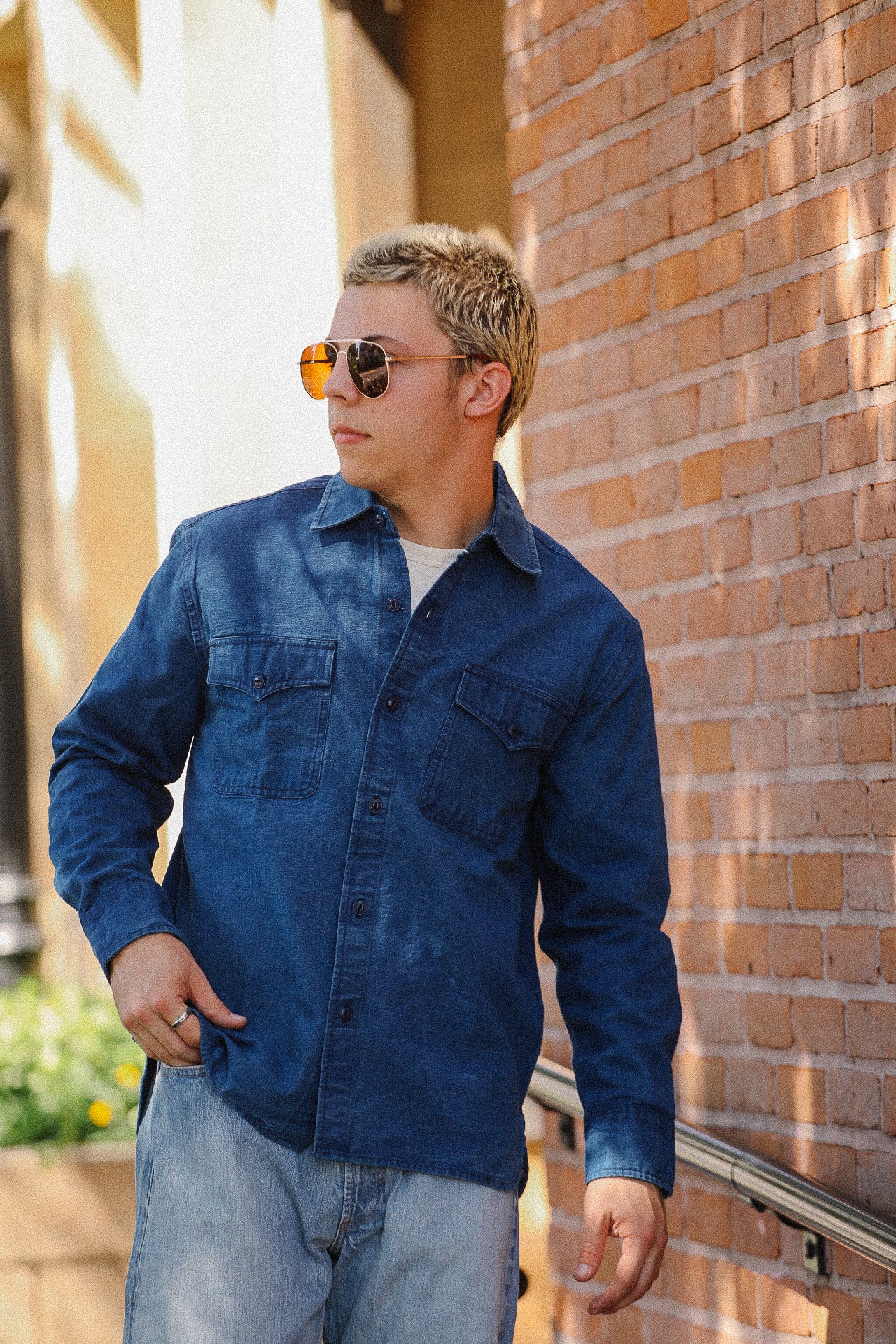 Man wearing blue denim shirt and aviator sunglasses standing against a brick wall outdoors