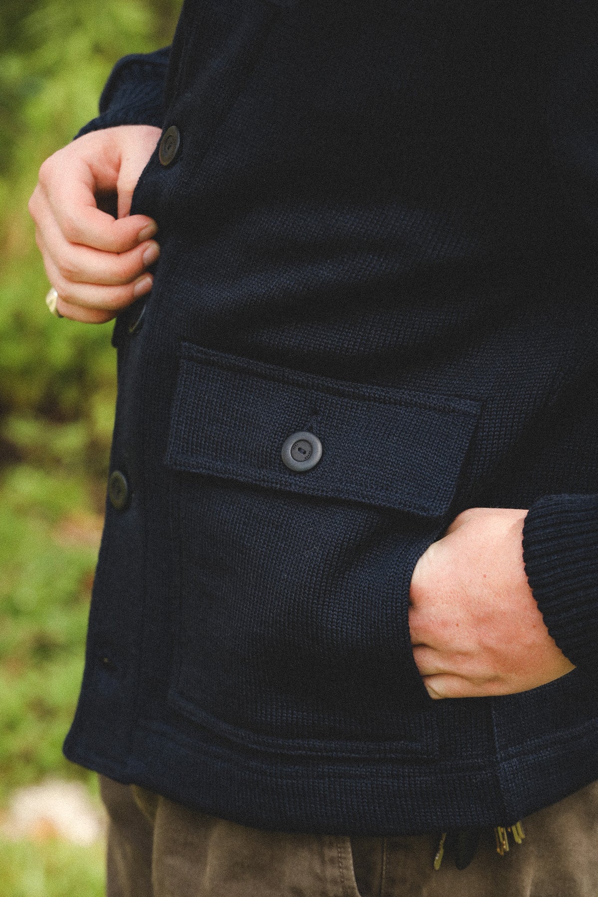 Close-up of a person wearing a dark navy textured knit cardigan with a buttoned chest pocket, outdoors with blurred green foliage in the background.