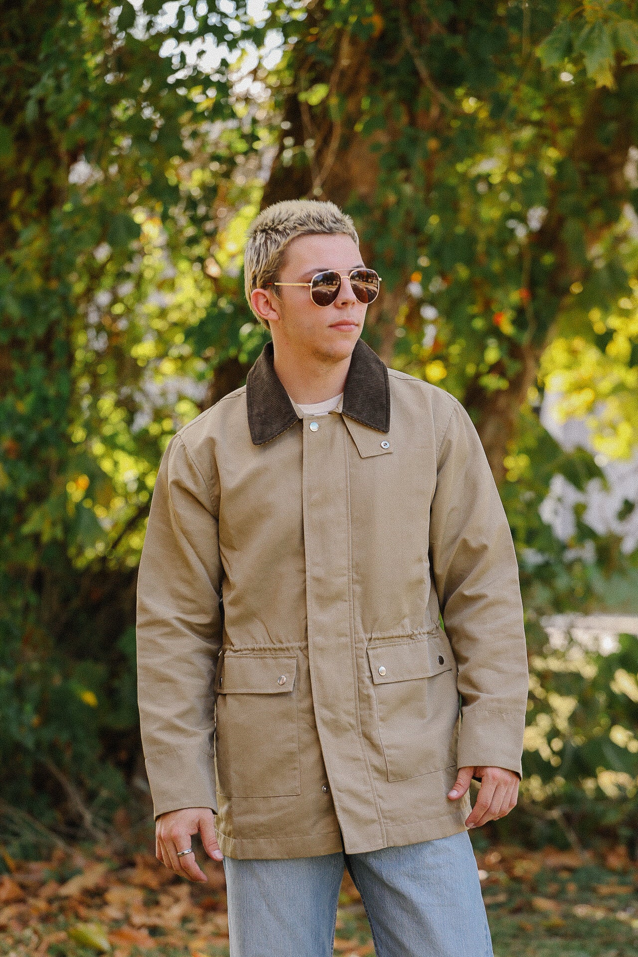 Man wearing a khaki canvas jacket with black collar, sunglasses, and light jeans outdoors