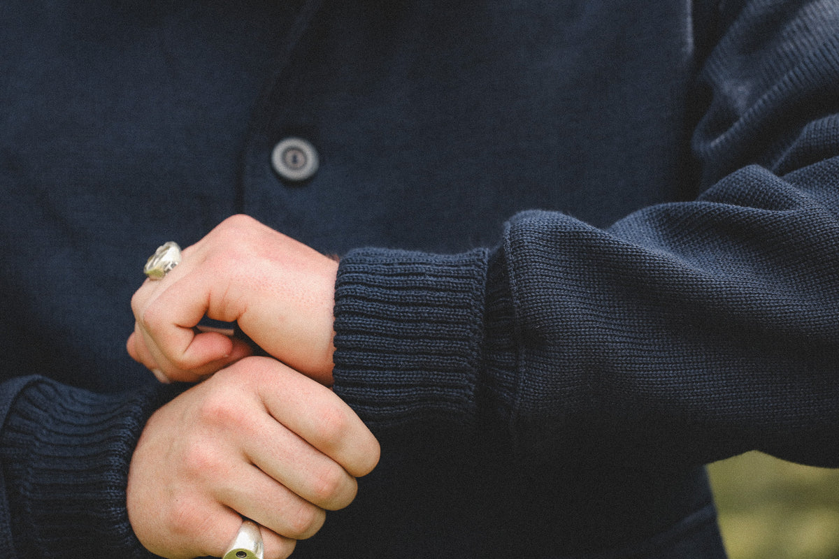 Close-up of a person wearing a dark navy ribbed knit cardigan sweater, with hands clasped in front and silver rings on the fingers.