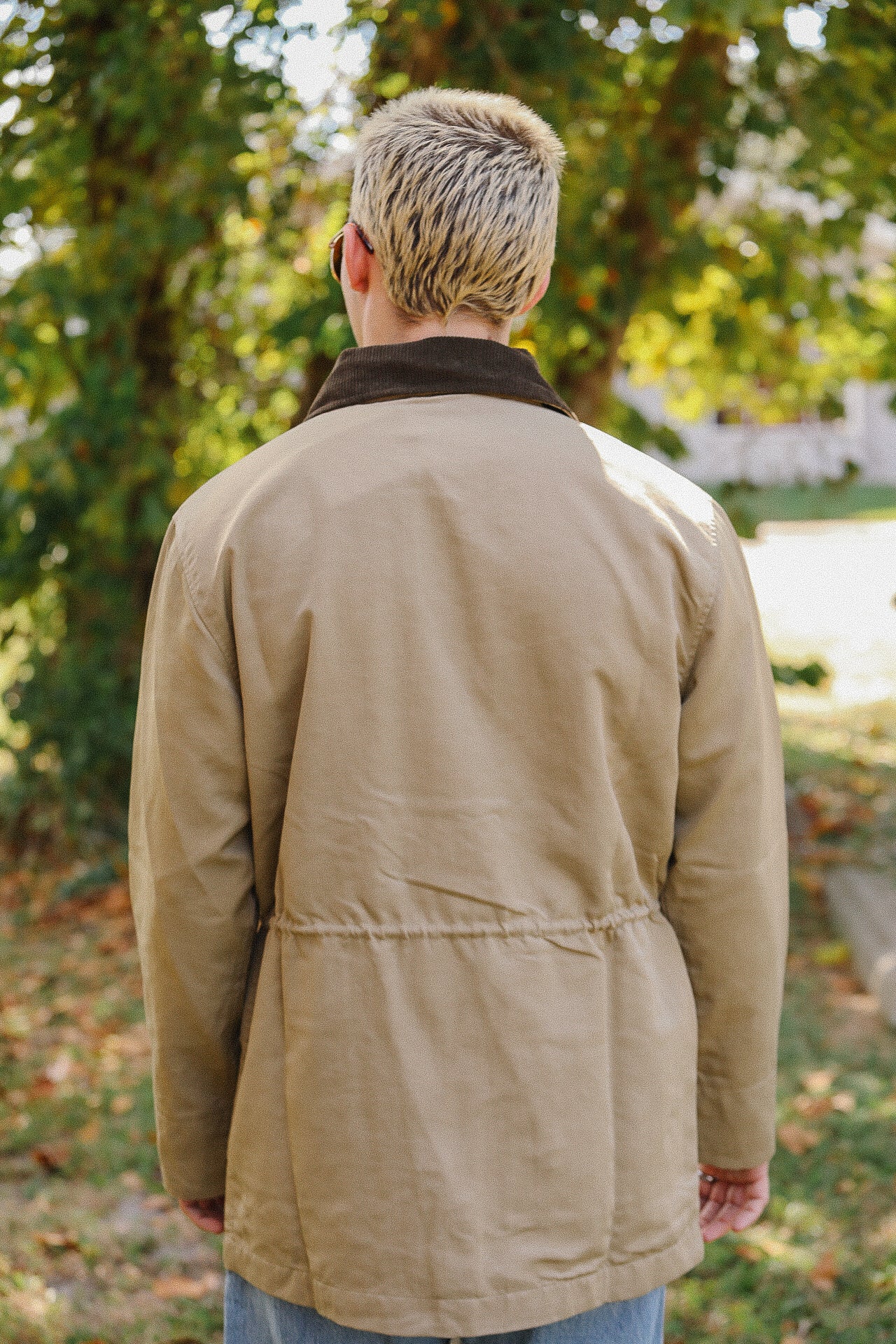 Person with short blond hair wearing tan jacket with dark collar standing outdoors near trees