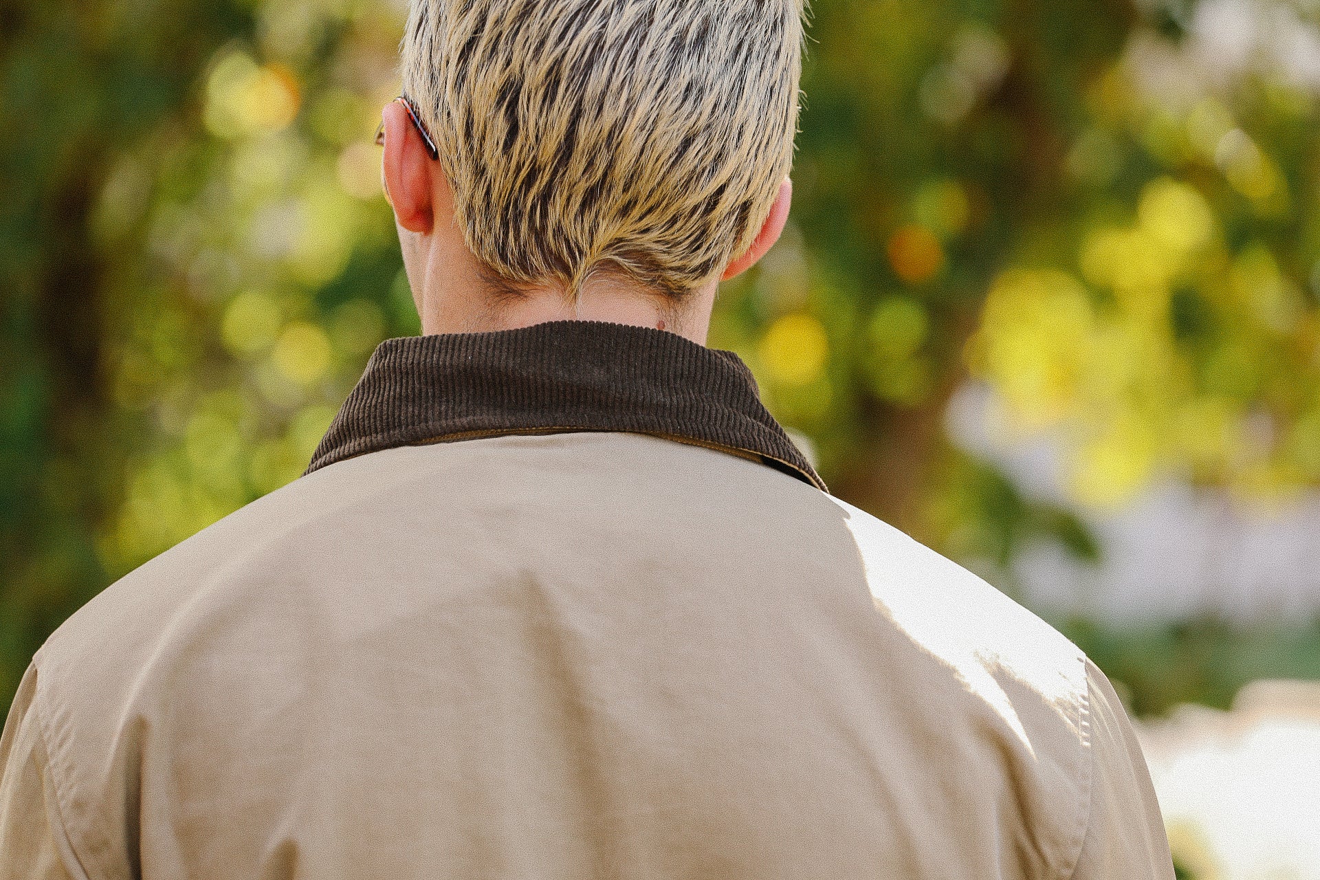 Close-up of a person with short blond hair wearing a beige jacket with dark brown corduroy collar in natural outdoor setting