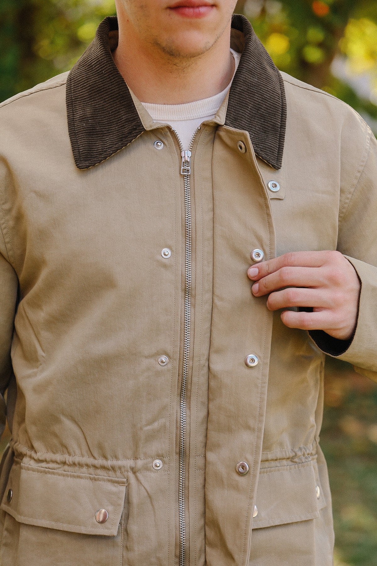Close-up of a person wearing a beige jacket with dark corduroy collar and metal snap buttons outdoors