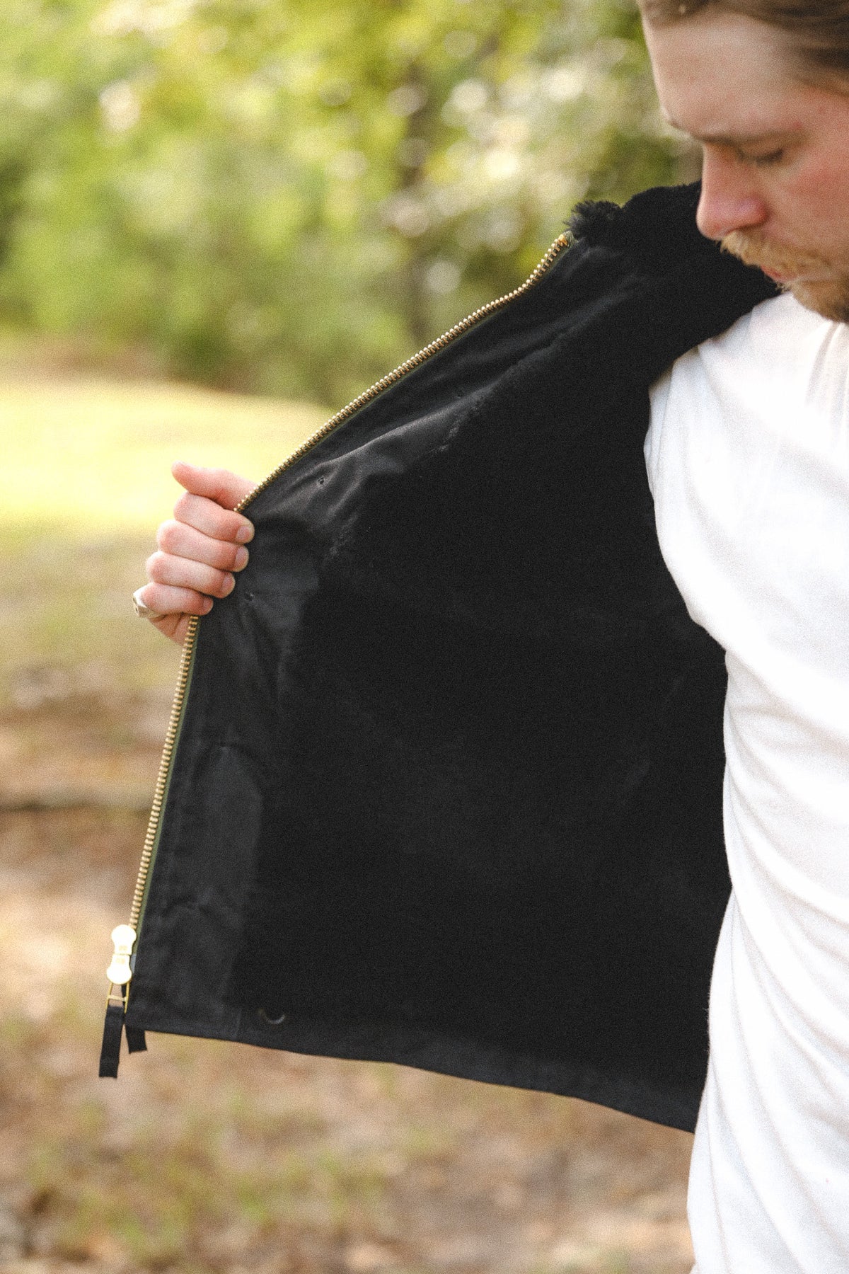 A man wearing a black zip-up jacket with a black lining, holding the zipper open with his hand in an outdoor setting.