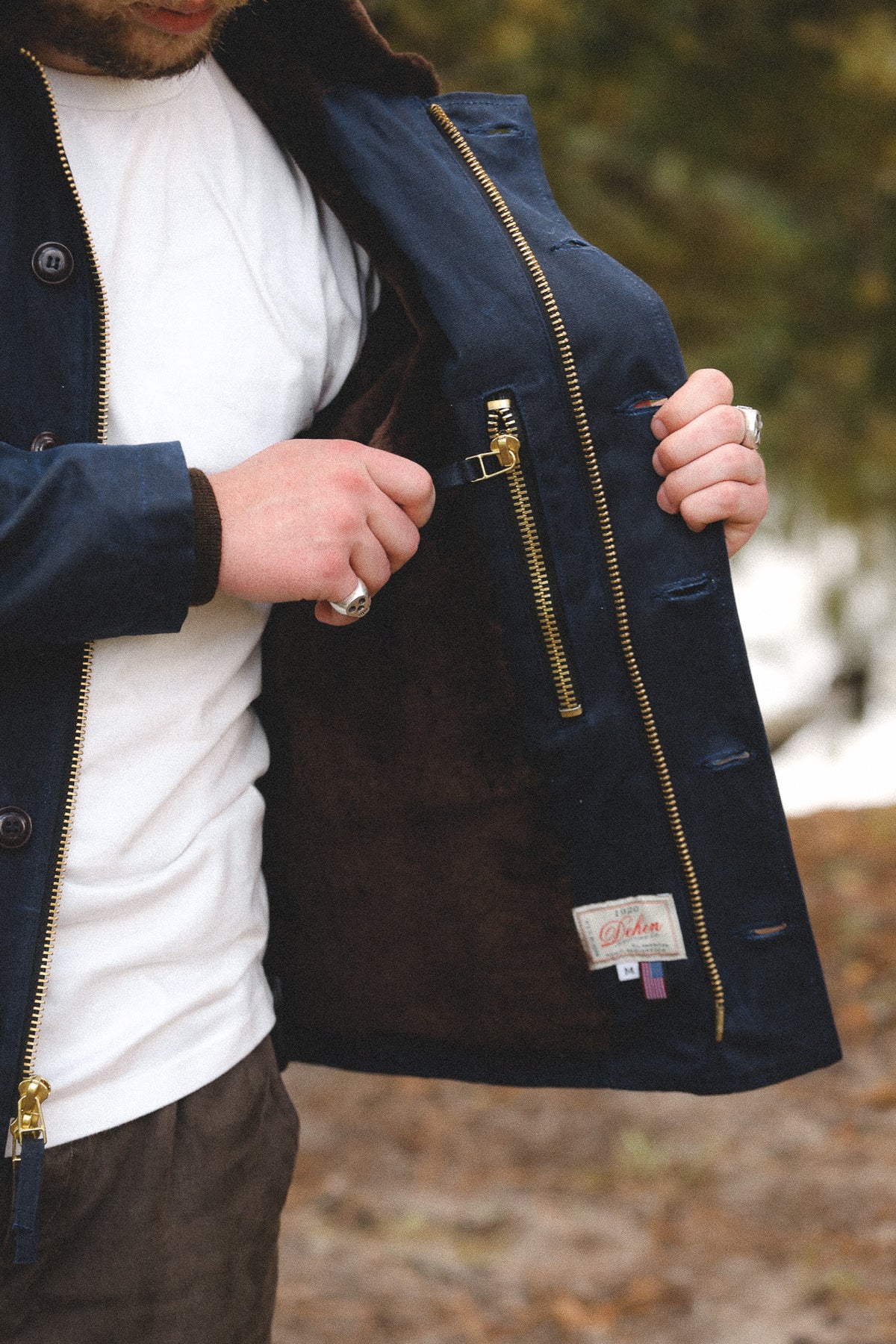 A close-up of a man wearing a navy blue jacket with a brown suede-like interior lining and gold-tone zippers, holding the jacket open to show the inner pocket. The background is a blurred outdoor setting.