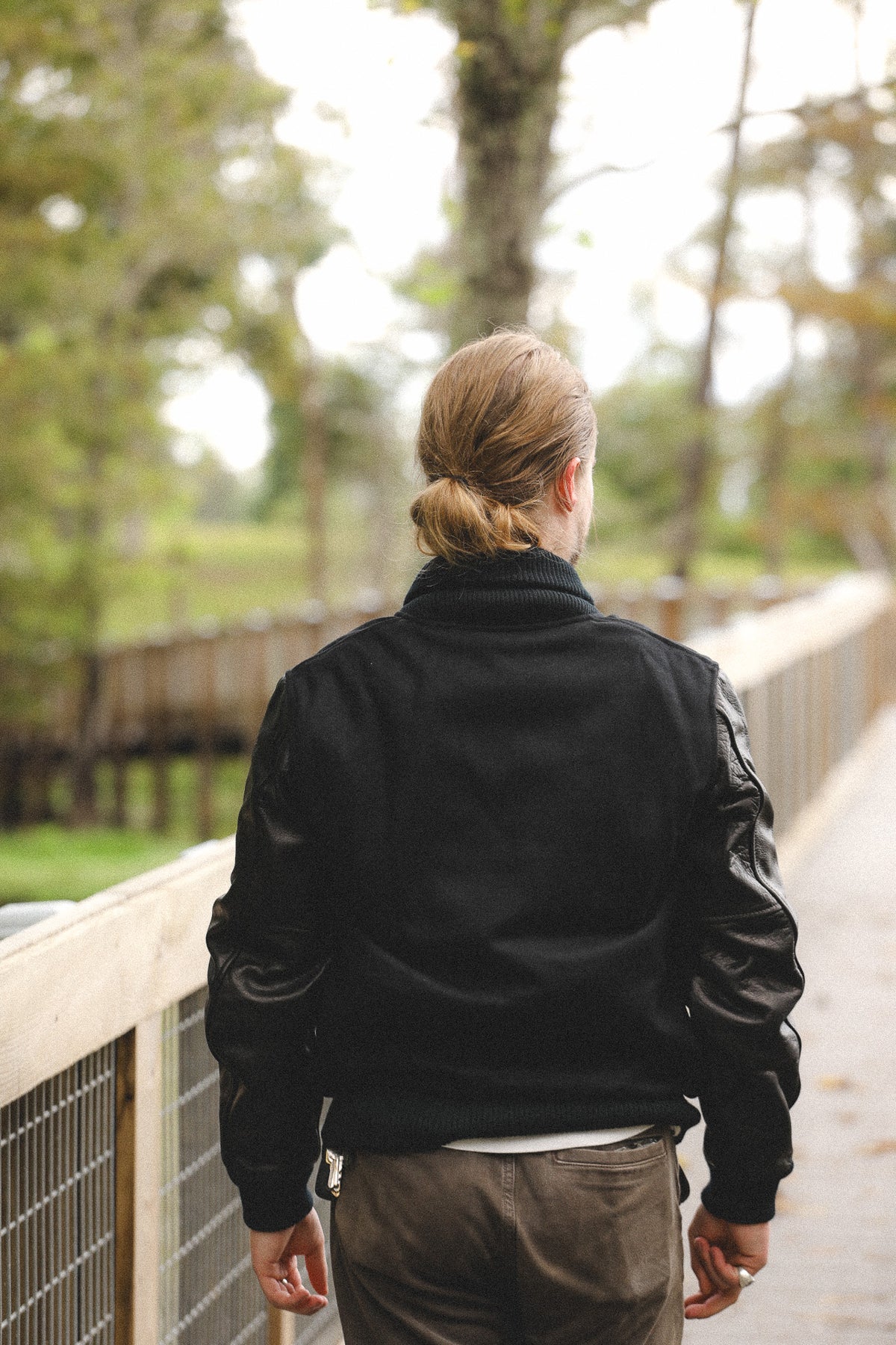 A person viewed from behind wearing a black bomber jacket with contrasting black leather sleeves, walking on an outdoor wooden boardwalk.