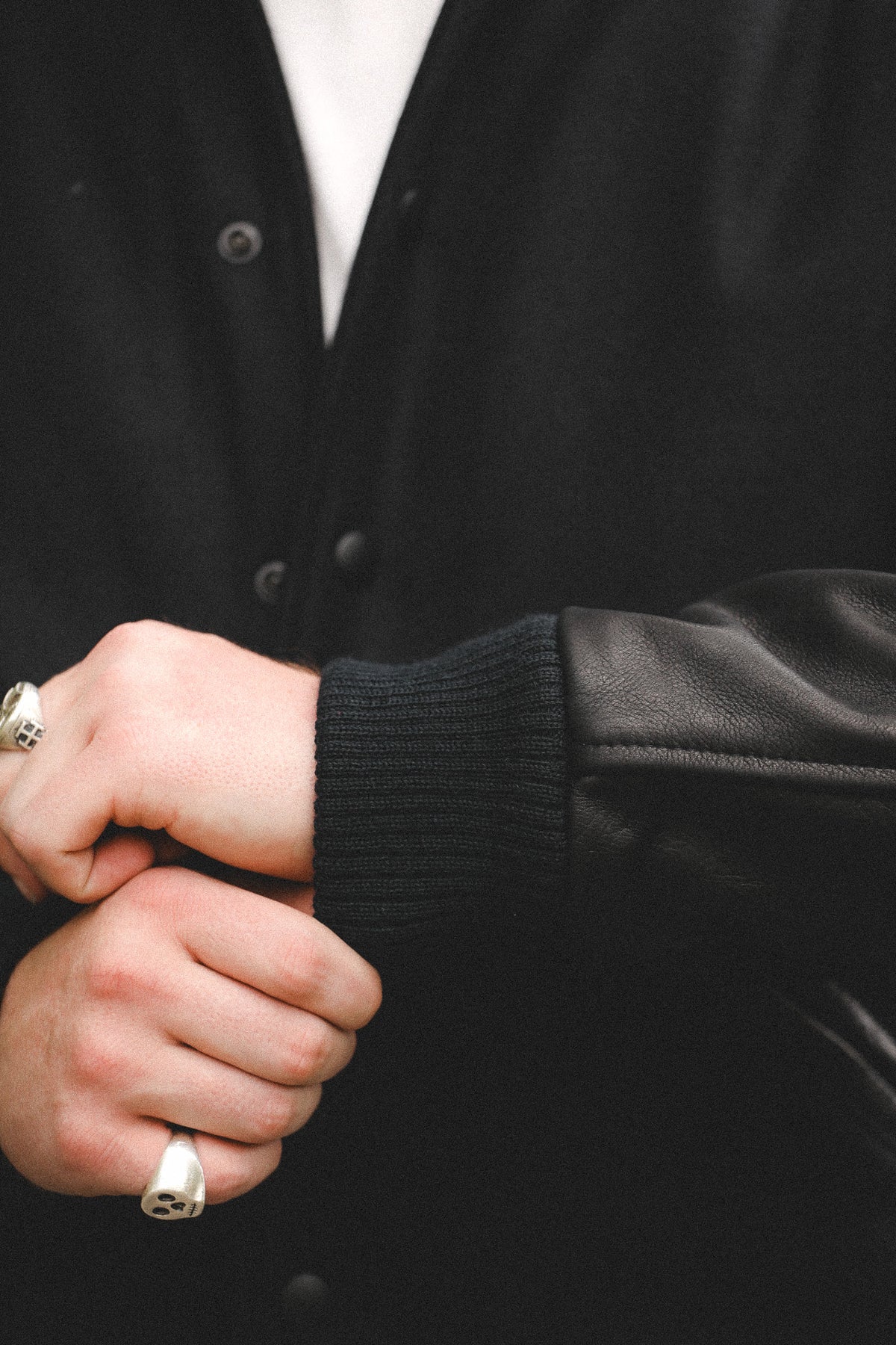 Close-up of a person wearing a black varsity-style jacket with a ribbed knit collar and a smooth leather sleeve, with silver rings on their fingers.