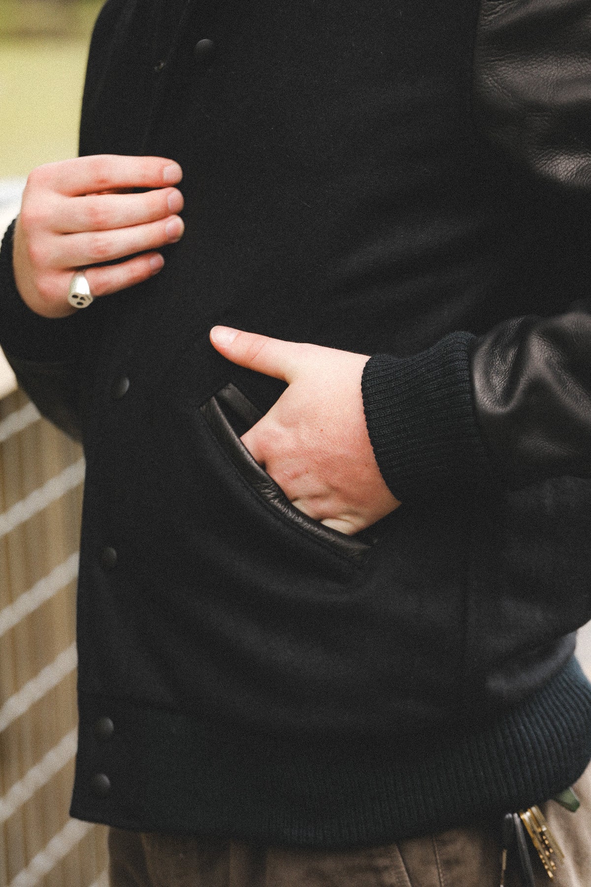 Close-up of a person wearing a black varsity-style jacket with dark leather sleeves and a side pocket, with one hand in the pocket and another hand resting near the chest.