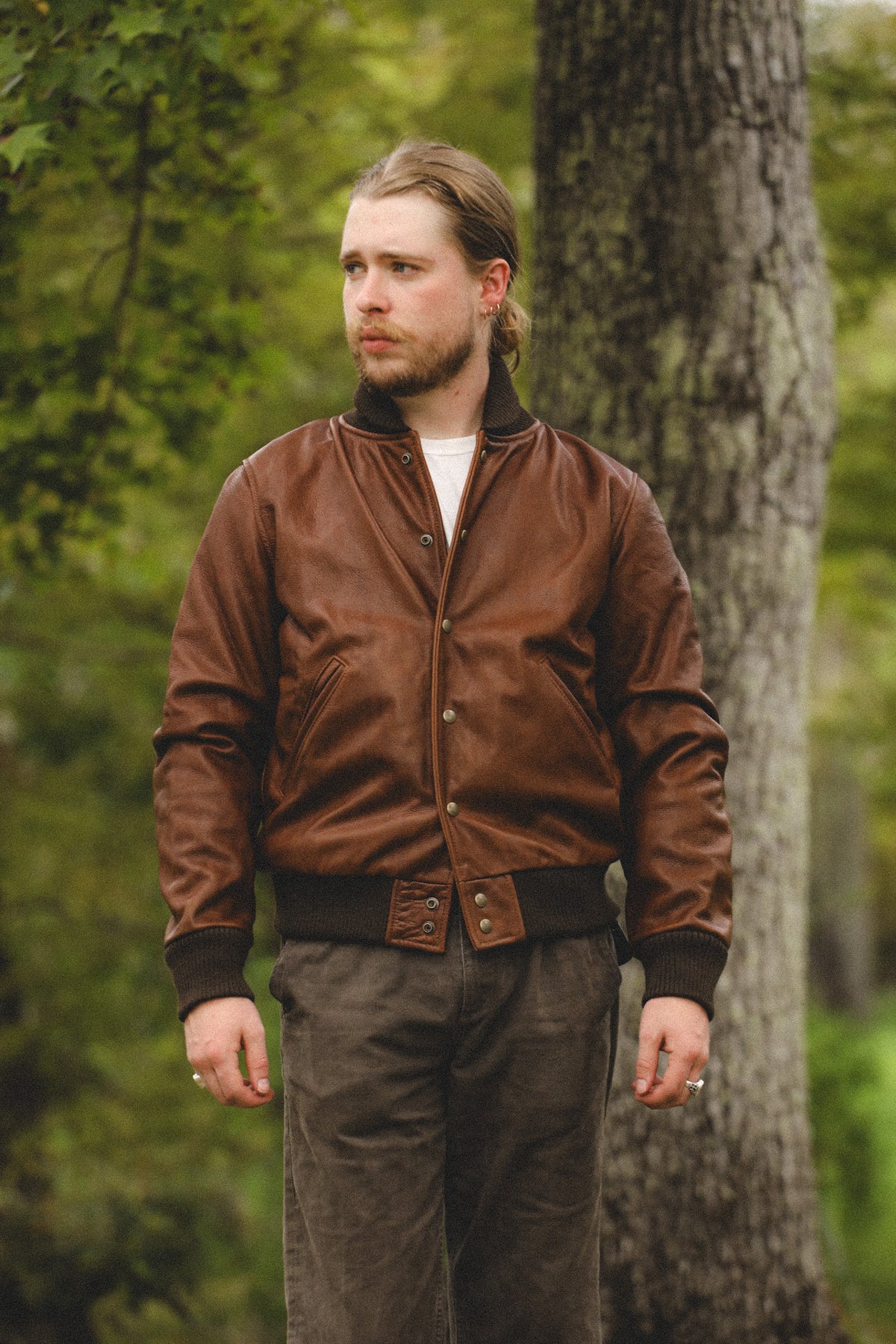A man wearing a brown leather bomber jacket with a black ribbed collar, cuffs, and hem, standing outdoors in front of a tree.