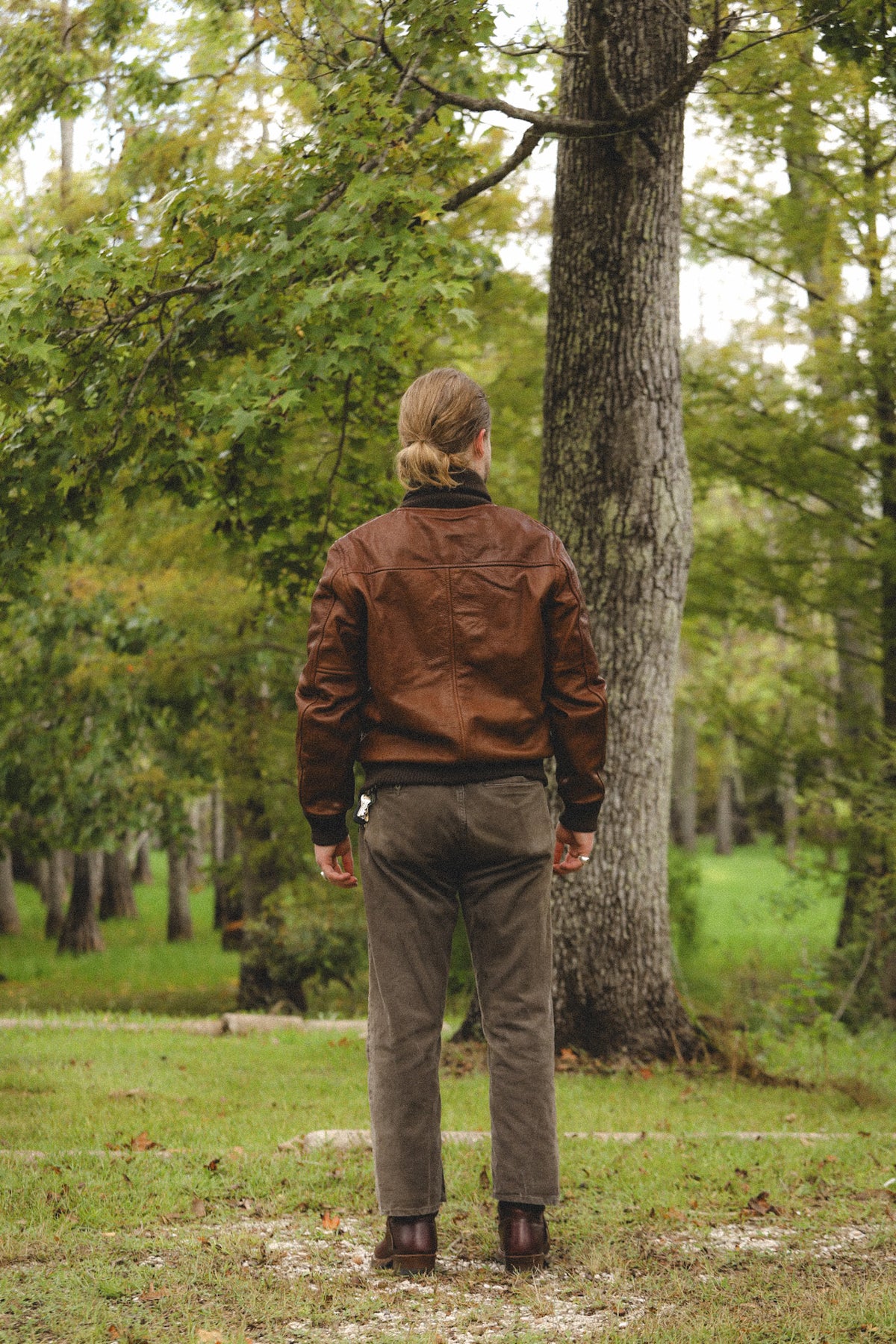 A man viewed from behind wearing a brown leather jacket, dark gray pants, and brown boots, standing in a grassy forest clearing next to a large tree trunk.