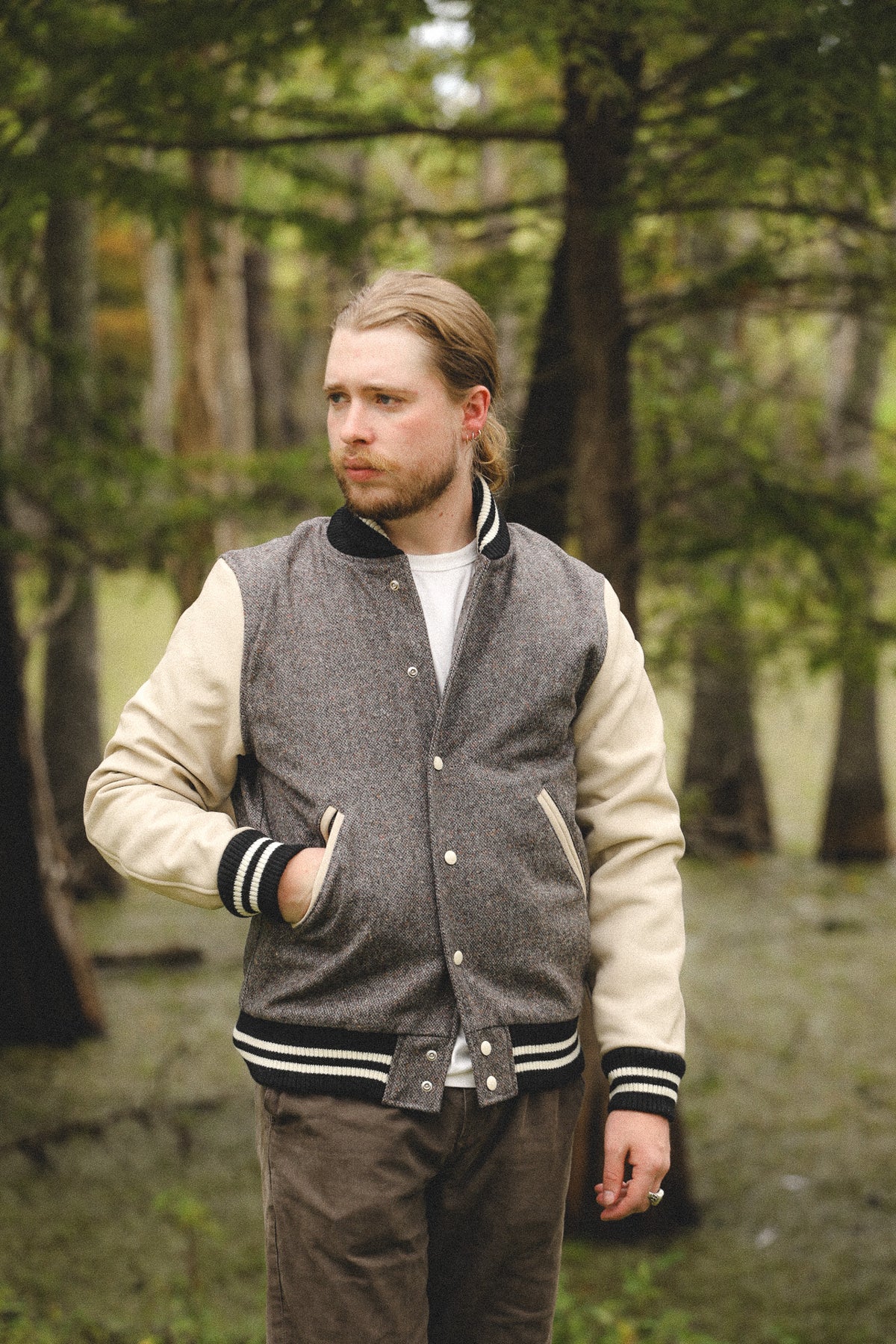 A man wearing a gray and cream varsity jacket standing in a forest.