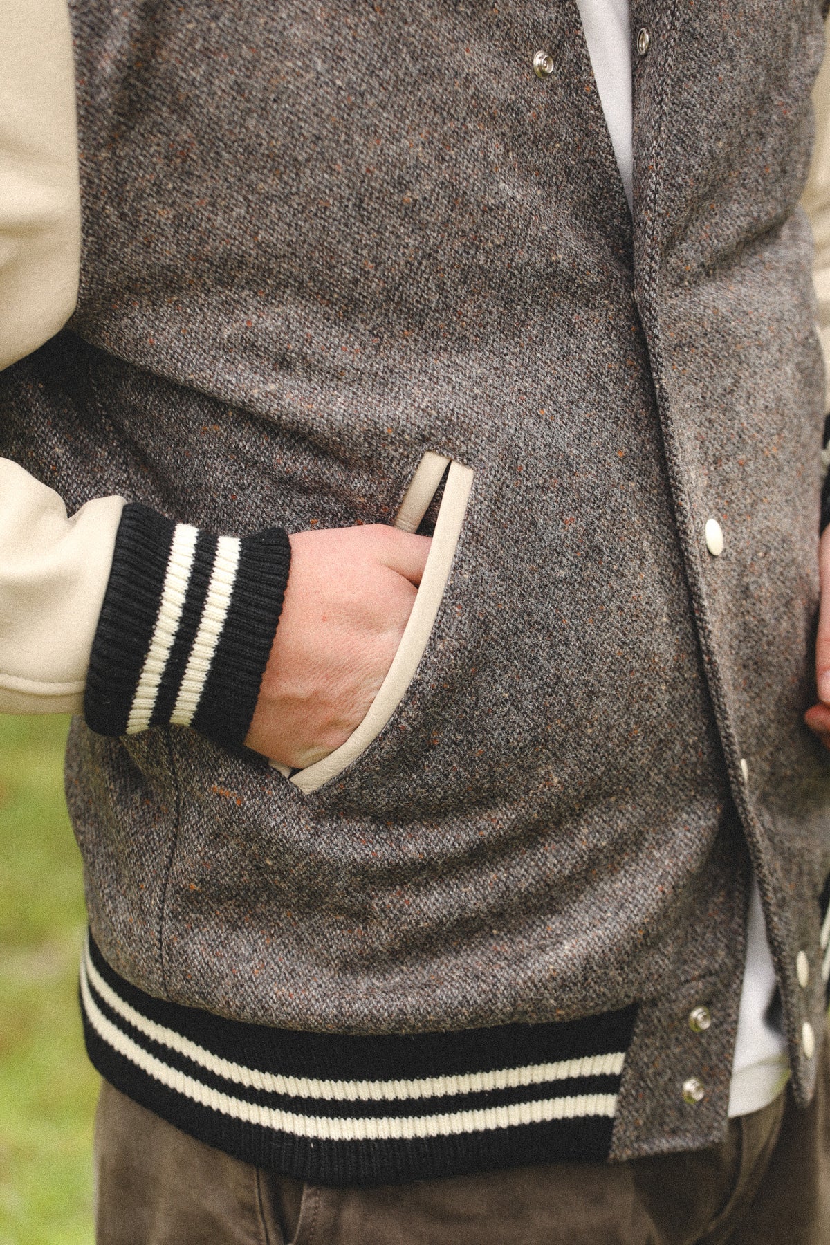 Close-up of a person wearing a gray and brown herringbone tweed varsity jacket with cream sleeves and black-and-white striped ribbed trim, with a hand in the pocket.