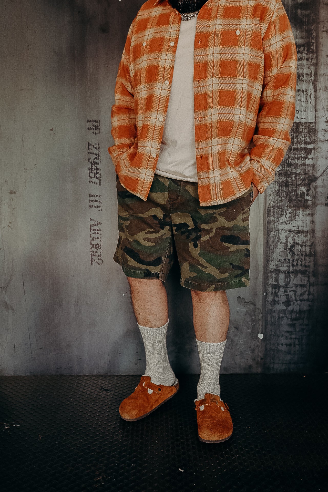 Man wearing orange plaid shirt, white T-shirt, camouflage shorts, gray socks, and brown suede slip-on shoes indoors