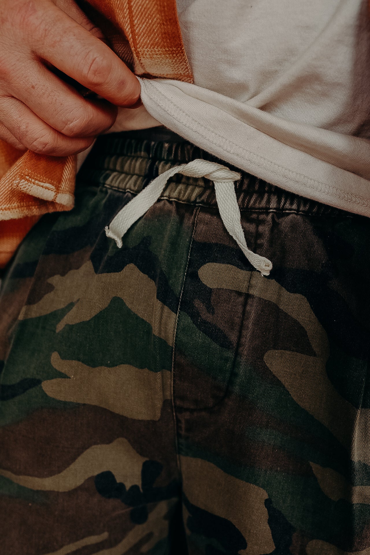 Close-up of man adjusting white tee and camo shorts with white drawstring at Iron Shop Provisions