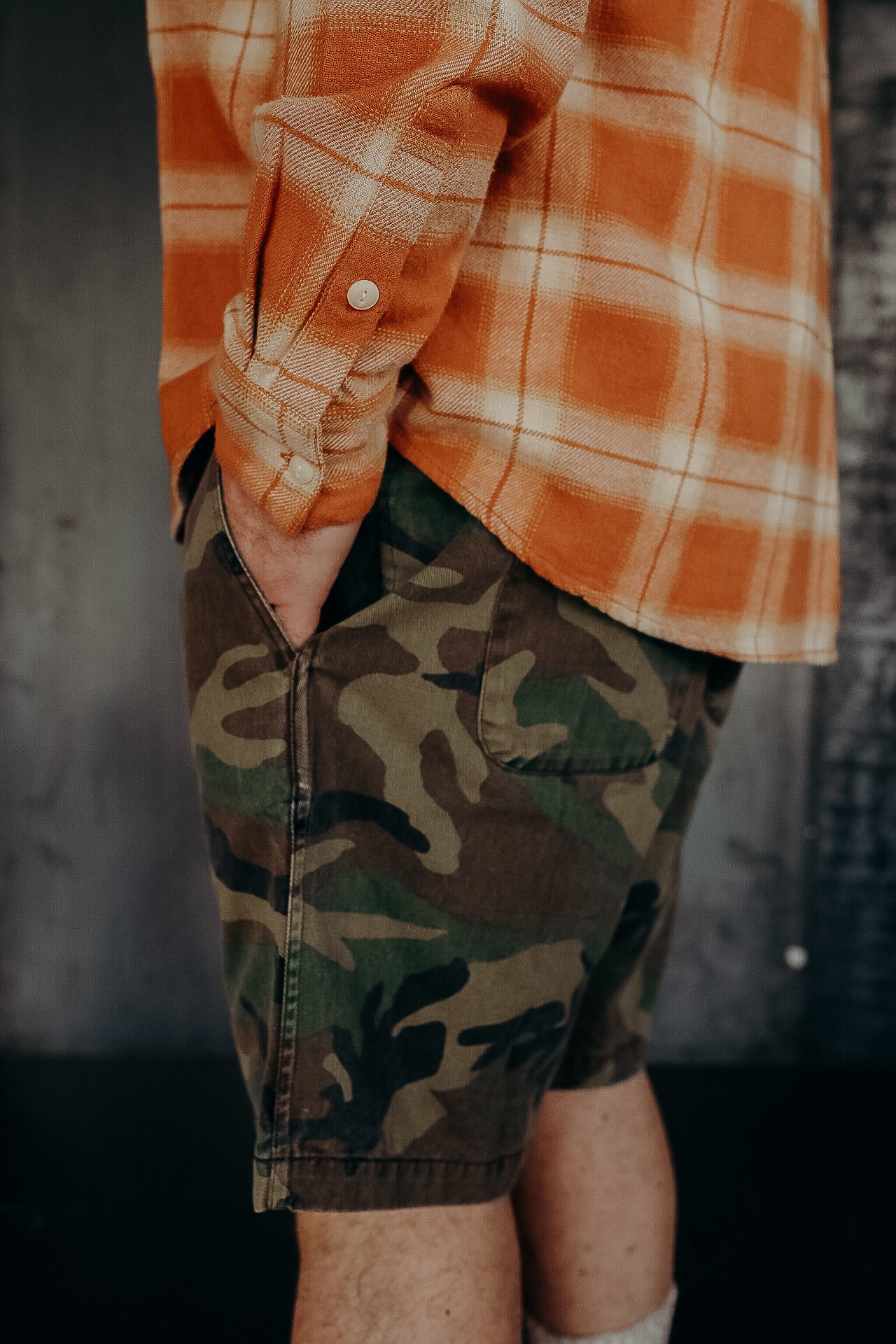 Man wearing orange plaid shirt and green camouflage shorts standing against a dark wall