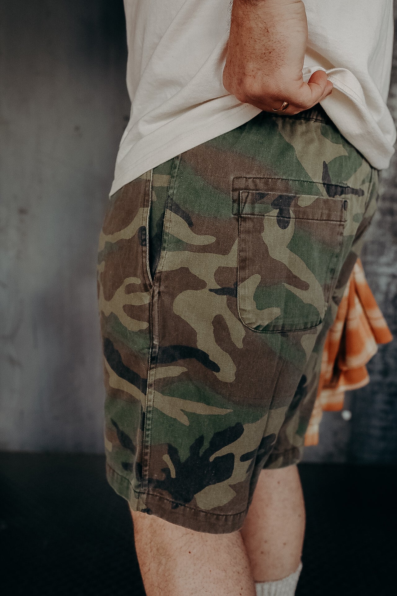 Close-up of person wearing green and brown camouflage shorts with back pocket, white shirt, and holding an orange plaid cloth