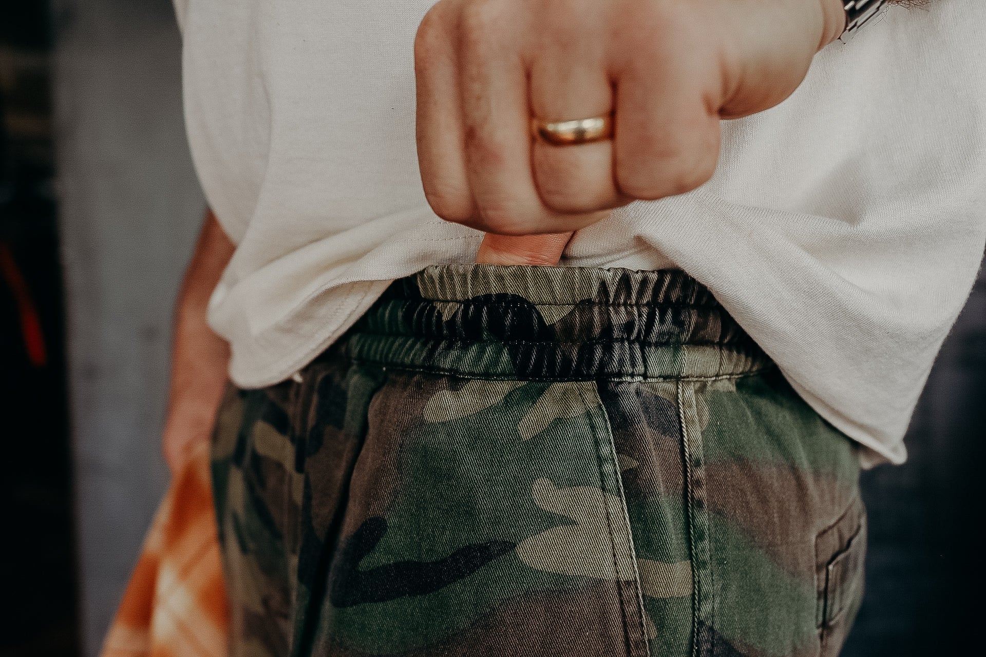 Close-up of a person wearing camo pants and a white shirt, hand tucked in pocket with ring visible