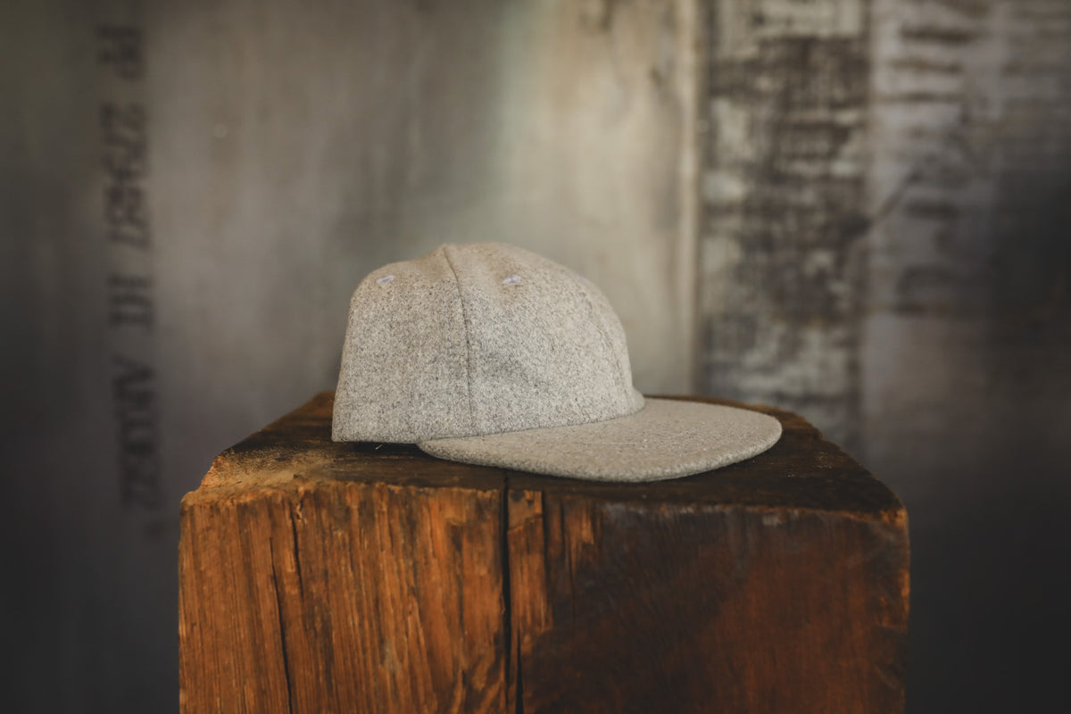 A light beige textured baseball cap with a flat visor, resting on a rustic wooden block against a blurred gray concrete background.