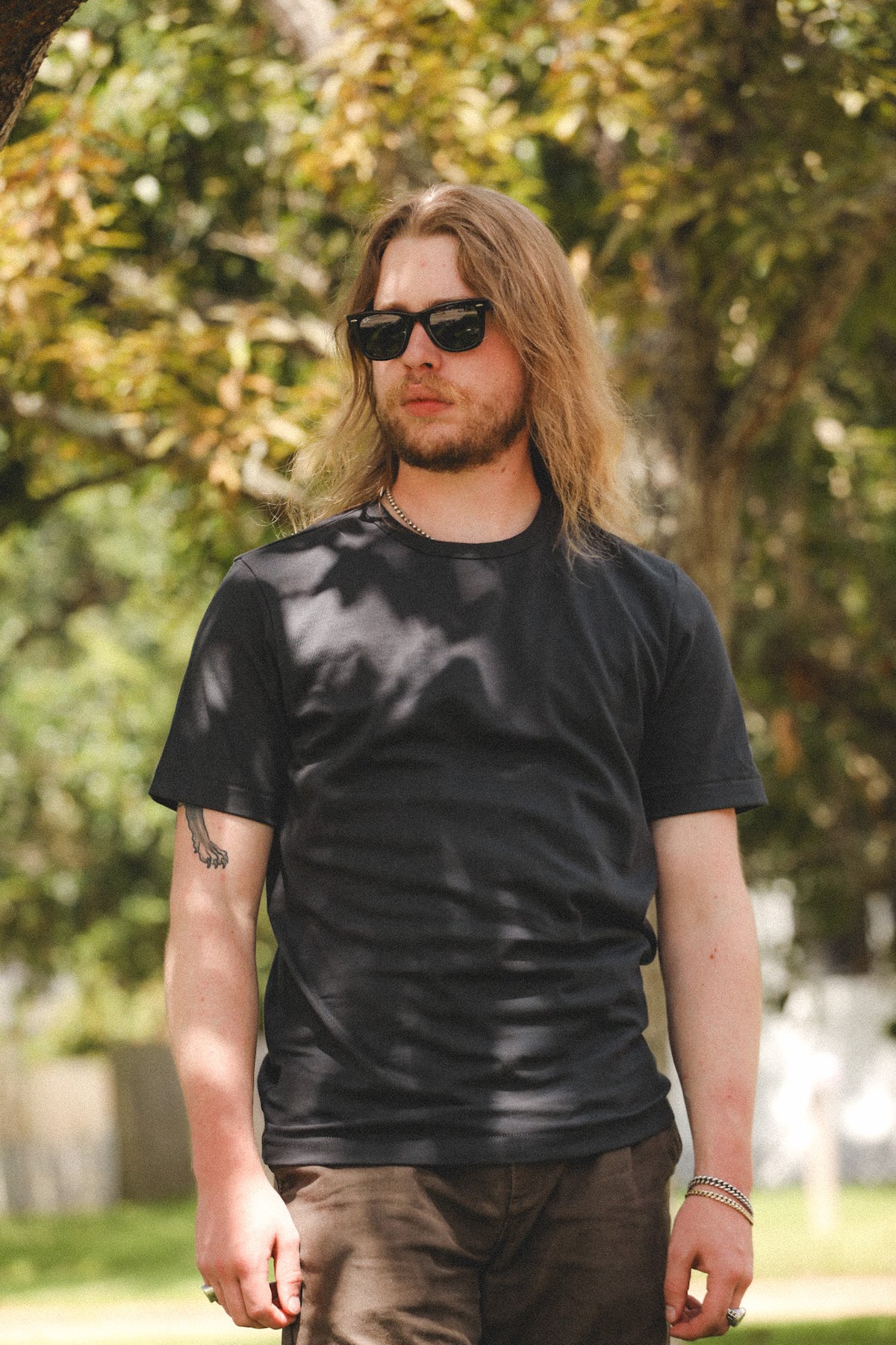 A man with long blonde hair and sunglasses wearing a black crew-neck t-shirt and brown pants, standing outdoors in front of a blurred background of green trees.