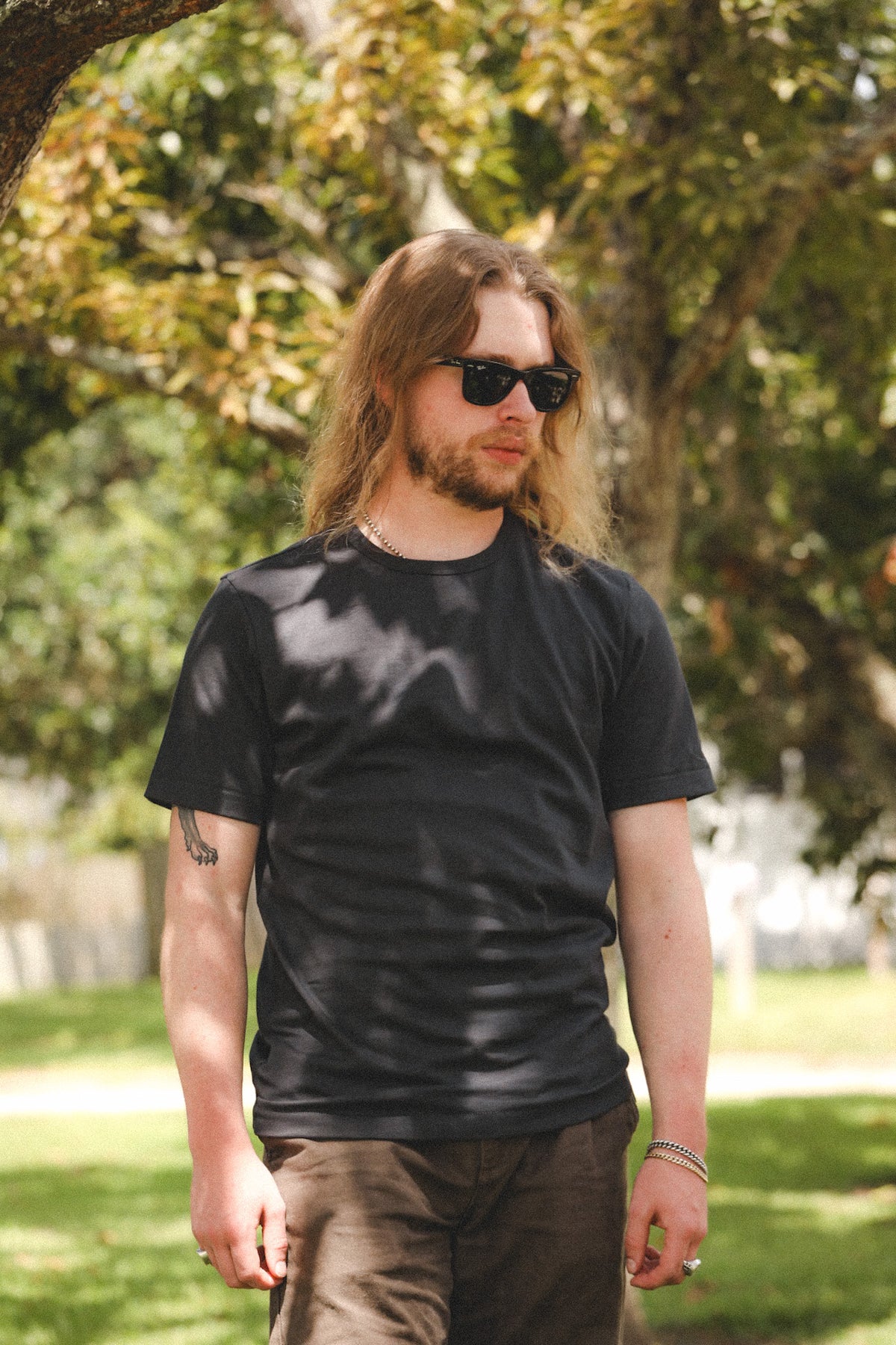 A man with long blonde hair, sunglasses, and a beard wearing a black crewneck t-shirt and brown pants, standing outdoors in a park with trees and grass in the background.