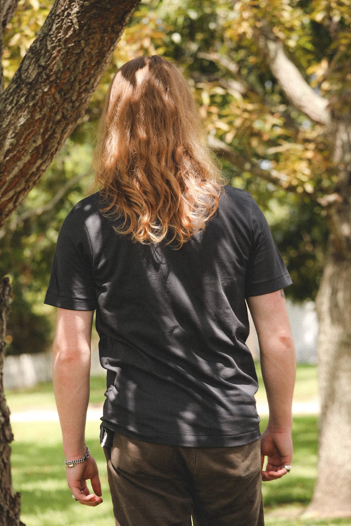 A person with long wavy light brown hair seen from behind, wearing a black short-sleeve t-shirt and brown pants, standing outdoors in a park with trees and grass.
