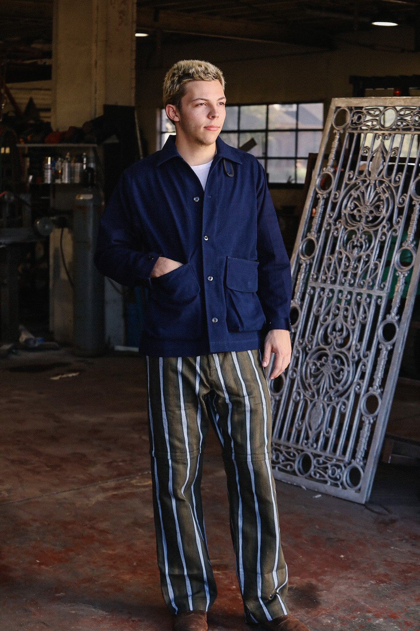 Man in navy blue work jacket and striped pants standing in a vintage industrial workshop with decorative iron gate