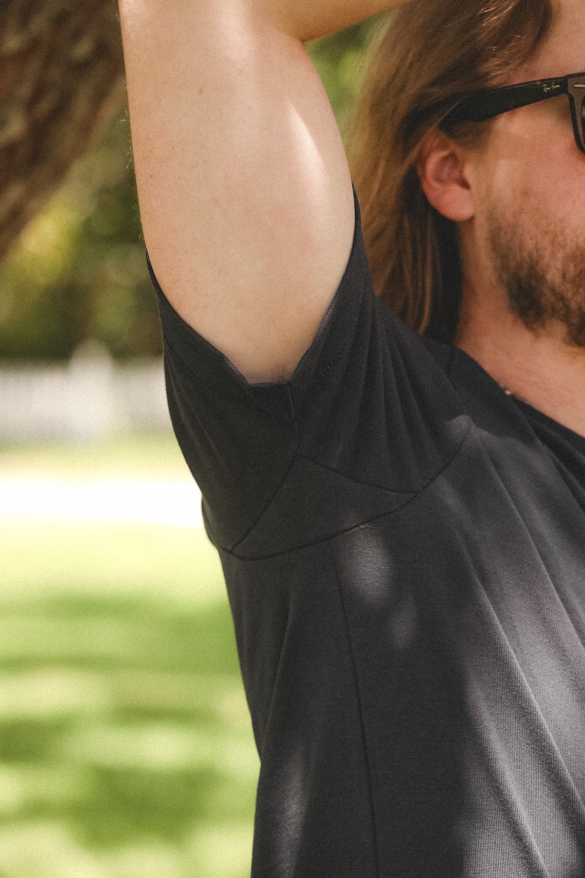 A close-up lifestyle shot of a man outdoors wearing a black athletic t-shirt with a high, structured shoulder and a wide armhole. He is wearing black sunglasses, and the background is a soft-focus green park.
