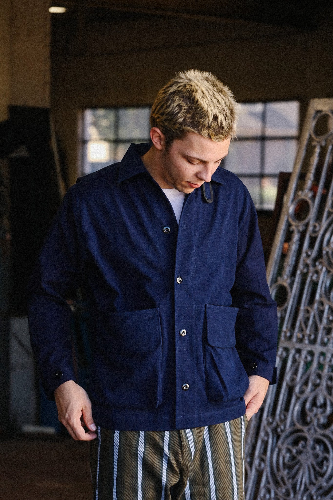 Man wearing navy work jacket with large pockets and striped pants in industrial workshop