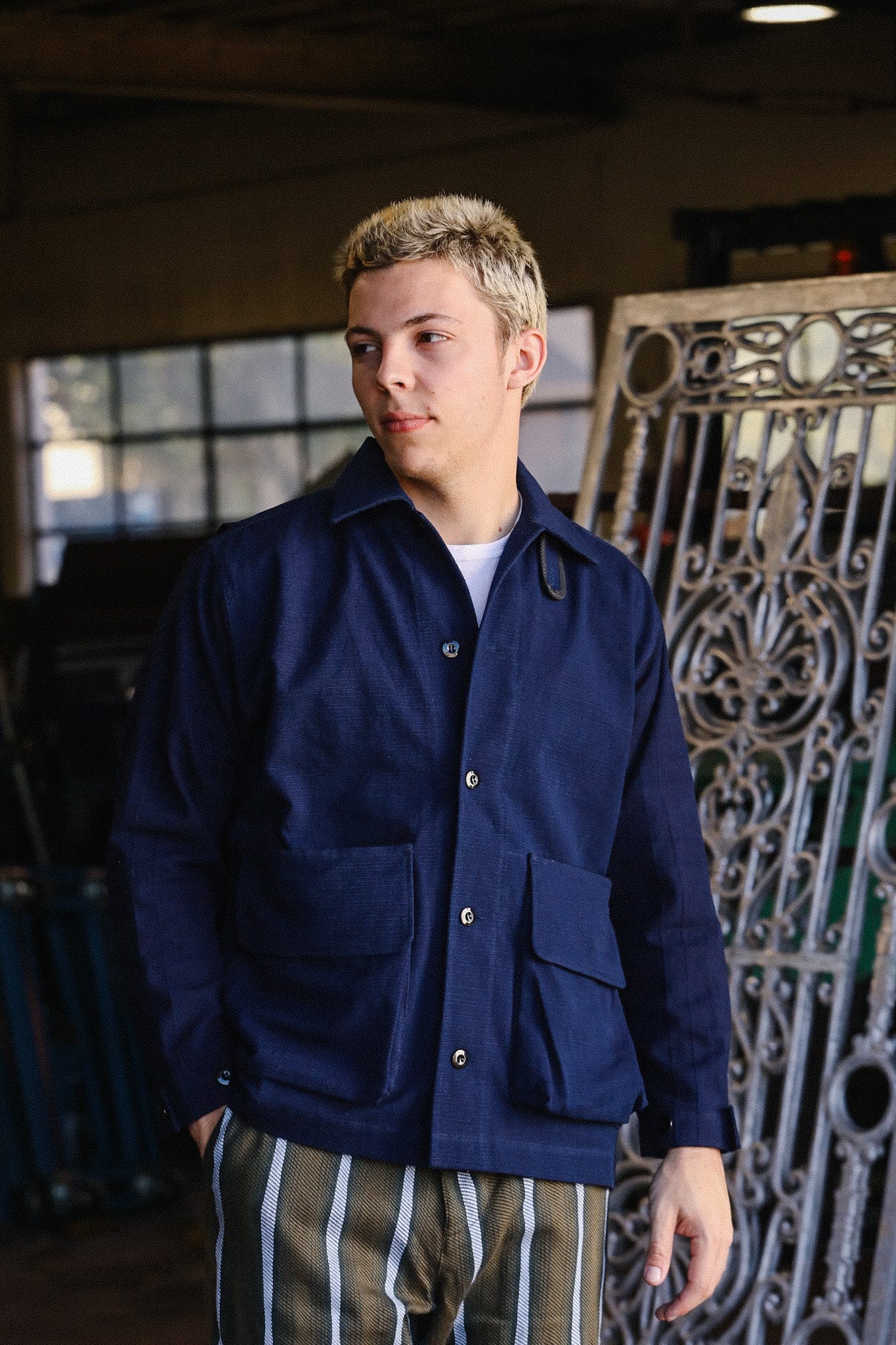 Young man in navy blue work jacket with large pockets and striped pants in industrial setting