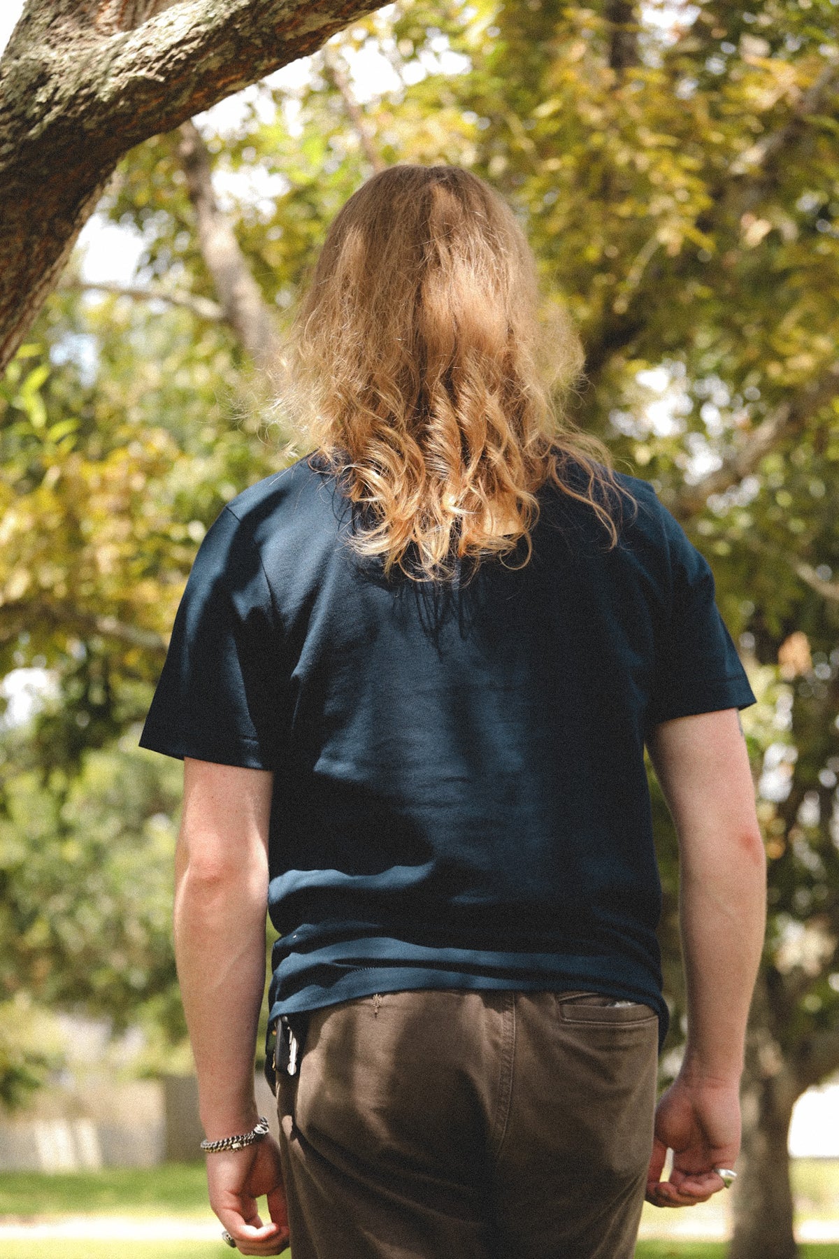 A person viewed from behind wearing a dark navy blue short-sleeve t-shirt and brown pants, standing outdoors in a park with trees and sunlight.