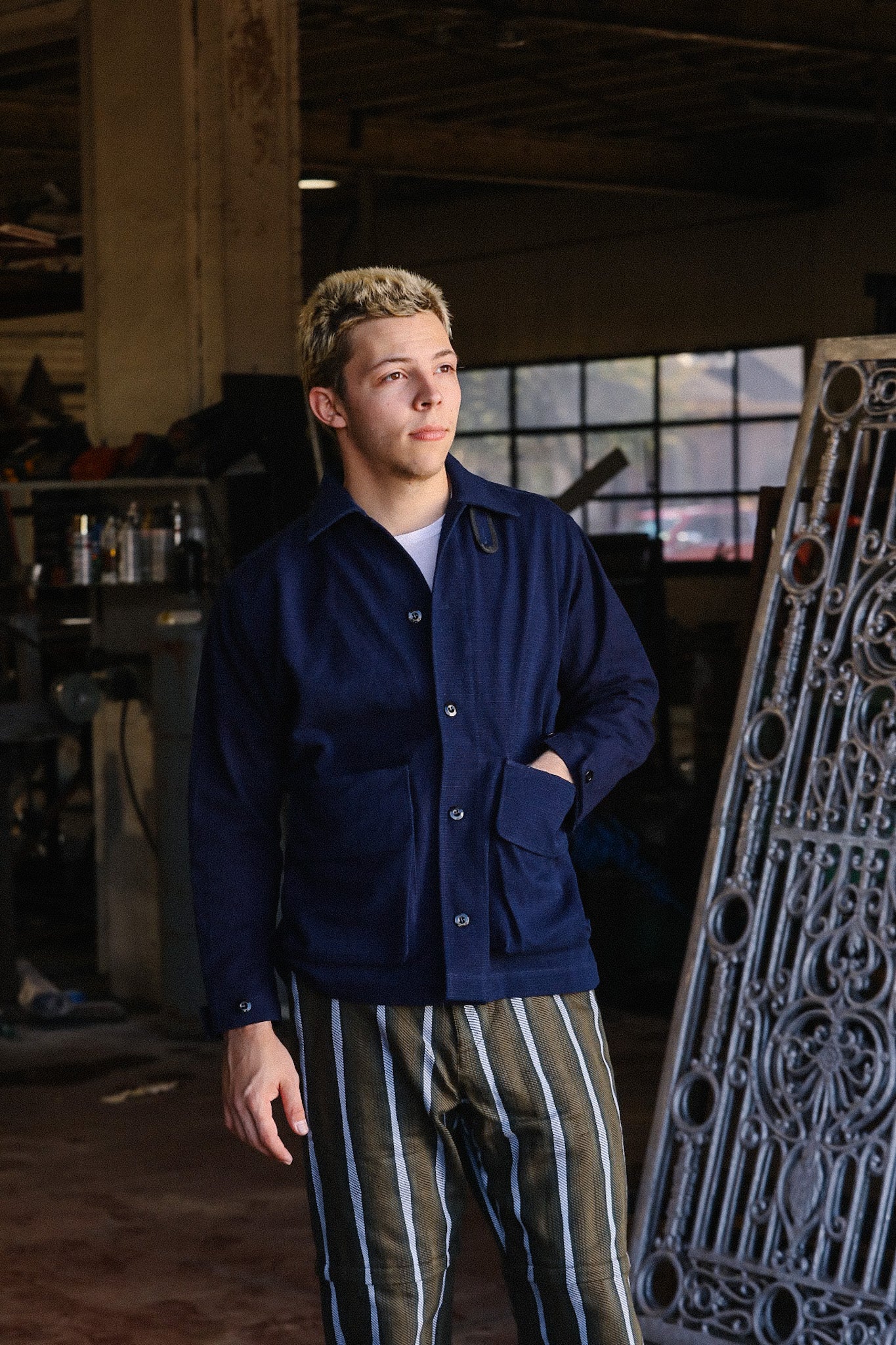 Young man in navy work jacket and striped pants posing in industrial workshop with decorative metal panel
