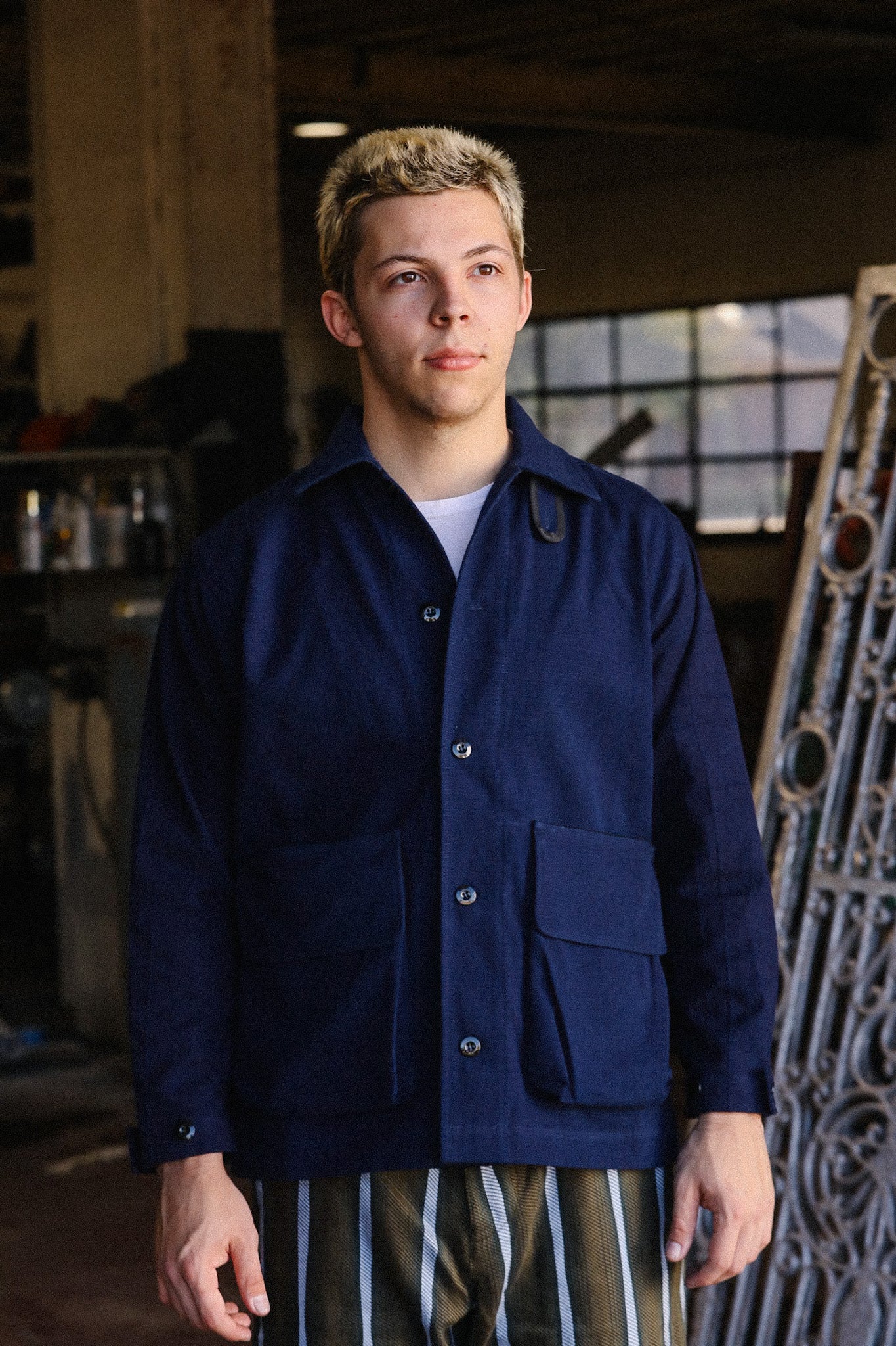 Young man wearing dark blue work jacket and striped pants in industrial workshop setting