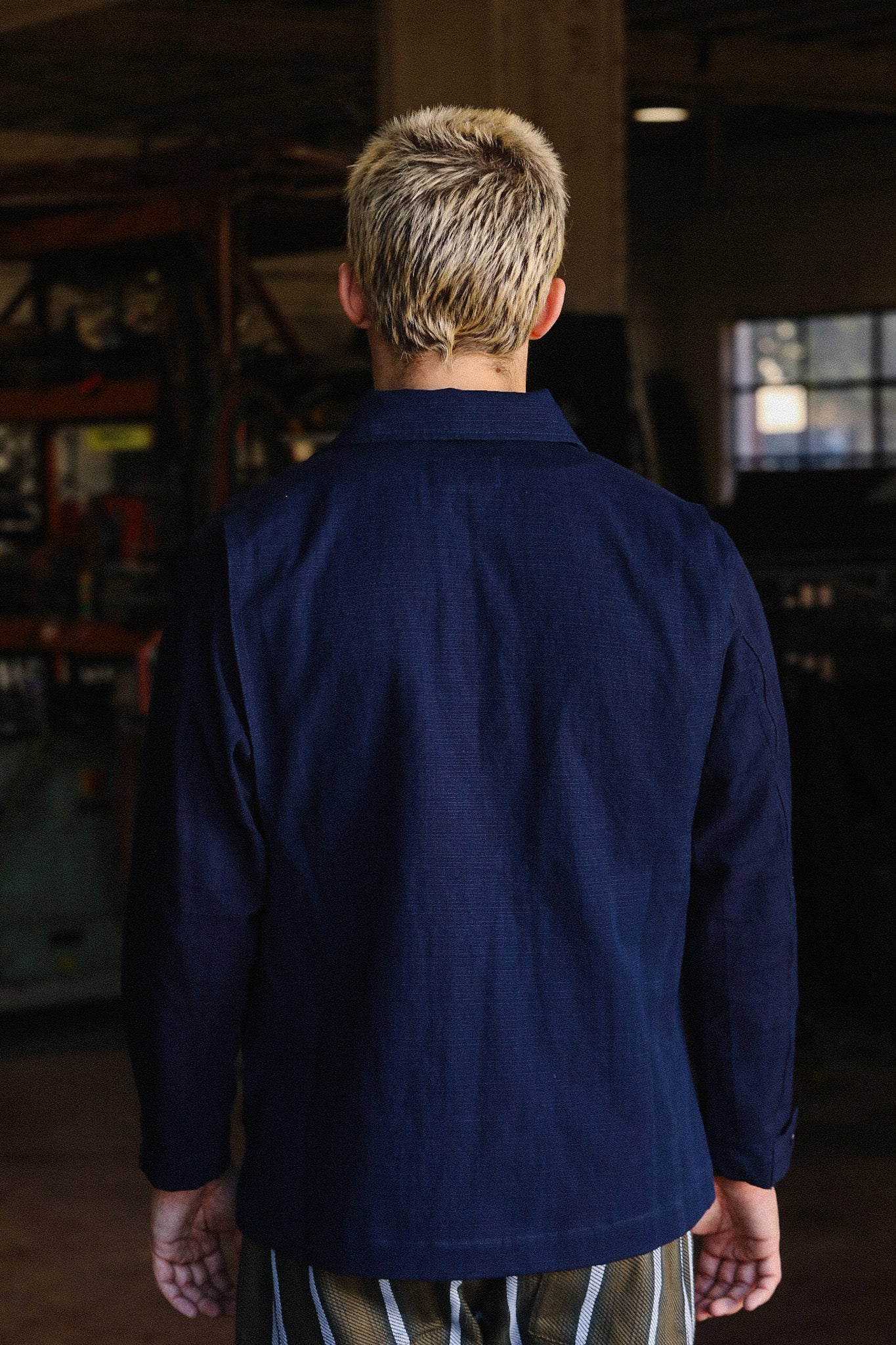 Person with short bleached hair wearing a navy blue jacket in an industrial workshop setting
