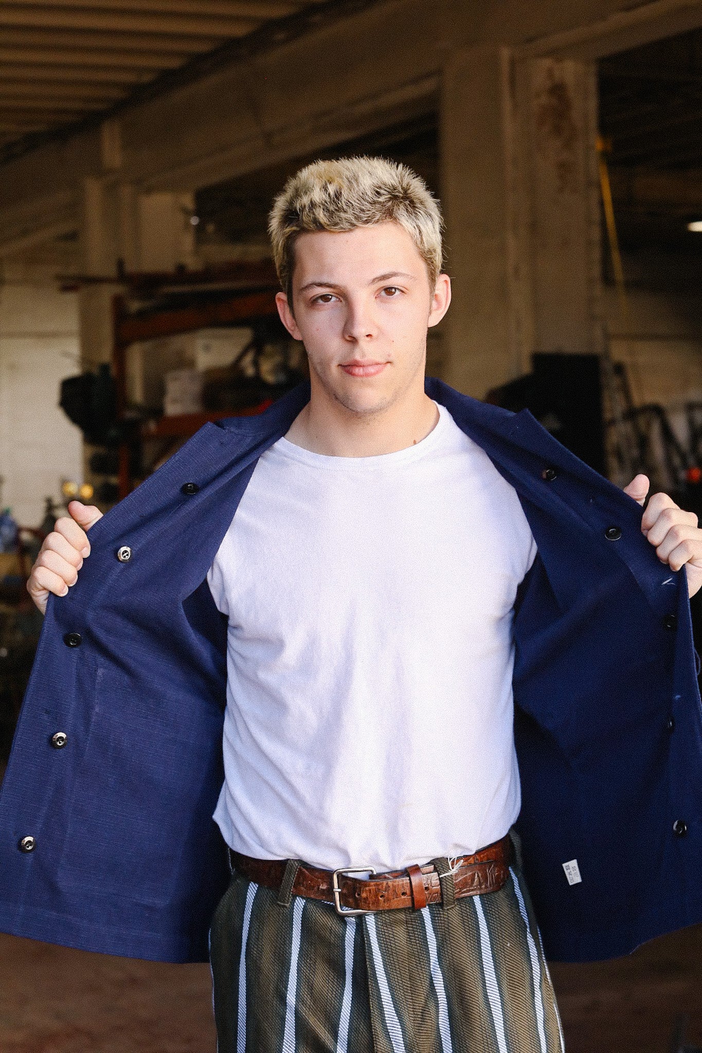 Young man in casual white t-shirt, striped pants, and blue jacket in industrial workshop setting