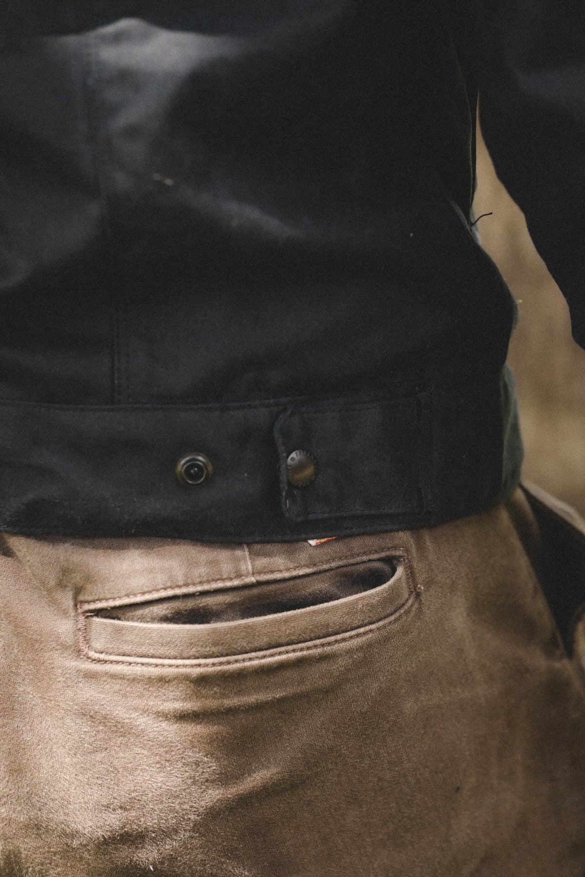 Close-up of a person wearing a black jacket with a buttoned flap at the waist and tan work pants, focusing on the back pocket area.