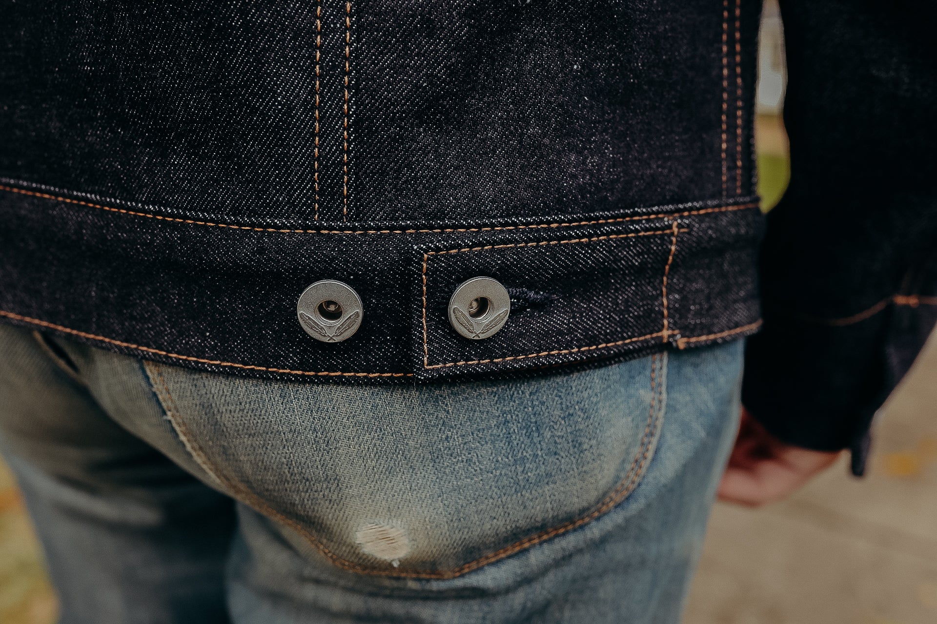Close-up of dark denim jacket hem with copper stitching and branded metal buttons over faded blue jeans