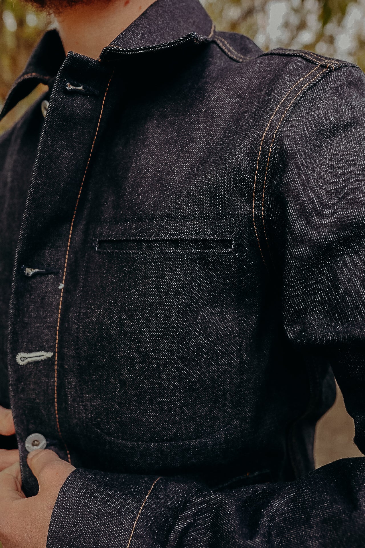 Close-up of person buttoning dark denim jacket with orange stitching from Iron Shop Provisions
