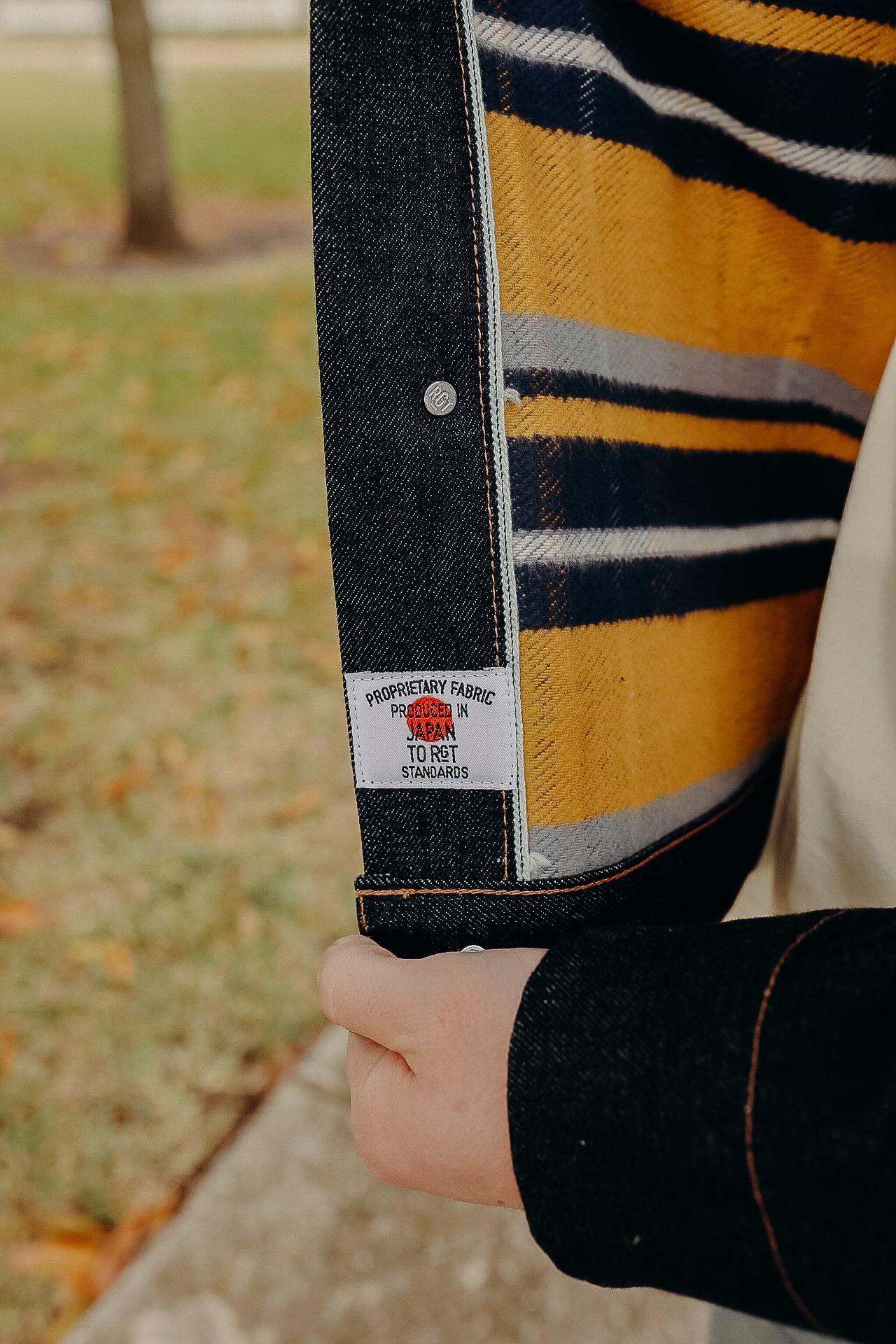 Person holding open a dark denim jacket showing its striped yellow and navy fabric lining and a label reading produced in Japan