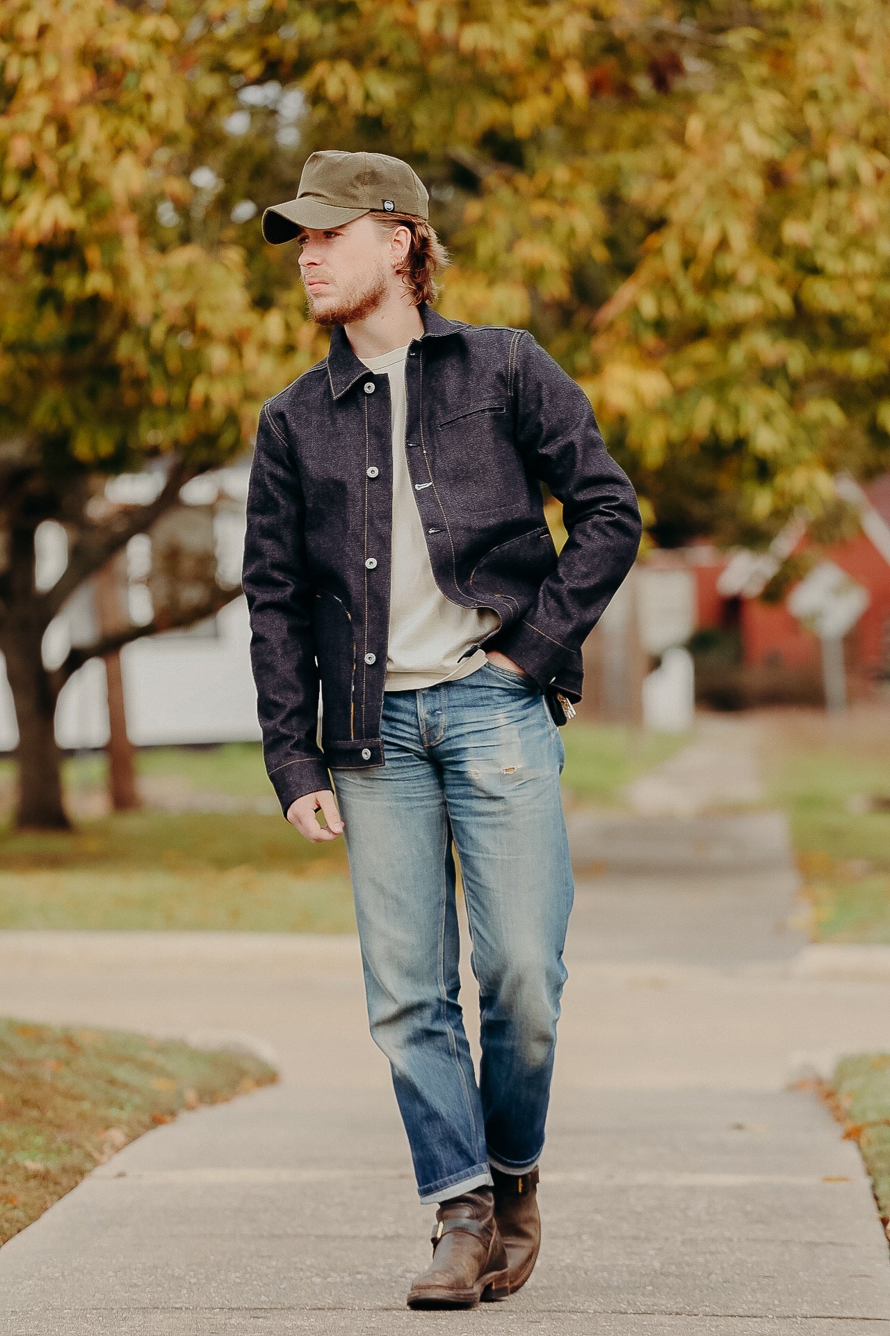 Man walking outdoors in fall wearing dark denim jacket, light shirt, blue jeans, brown boots, and olive cap