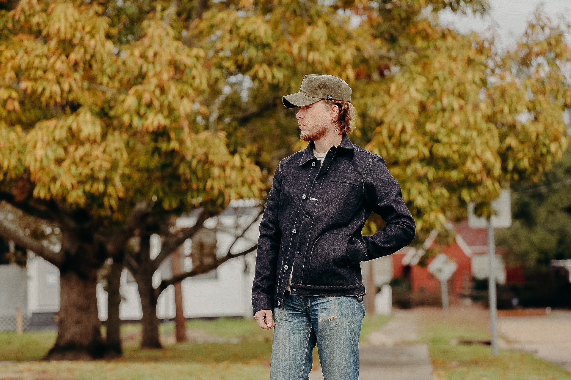 Man wearing dark denim jacket and green cap standing outdoors with autumn trees and houses in background