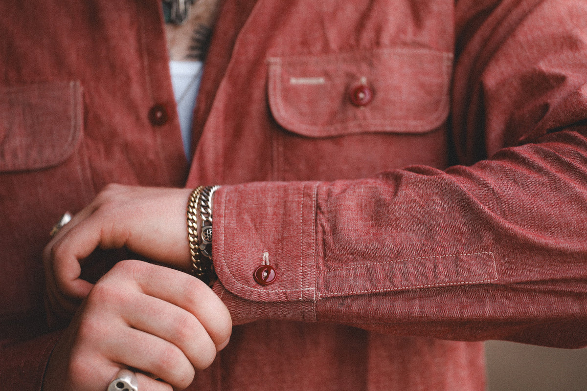 Close-up of a person wearing a rust-red corduroy button-up shirt jacket, adjusting the cuff with a gold chain bracelet on the wrist.