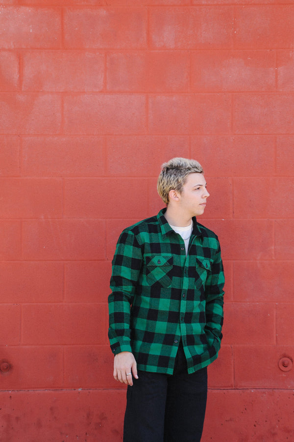 Young man in green and black checkered shirt standing against a red brick wall