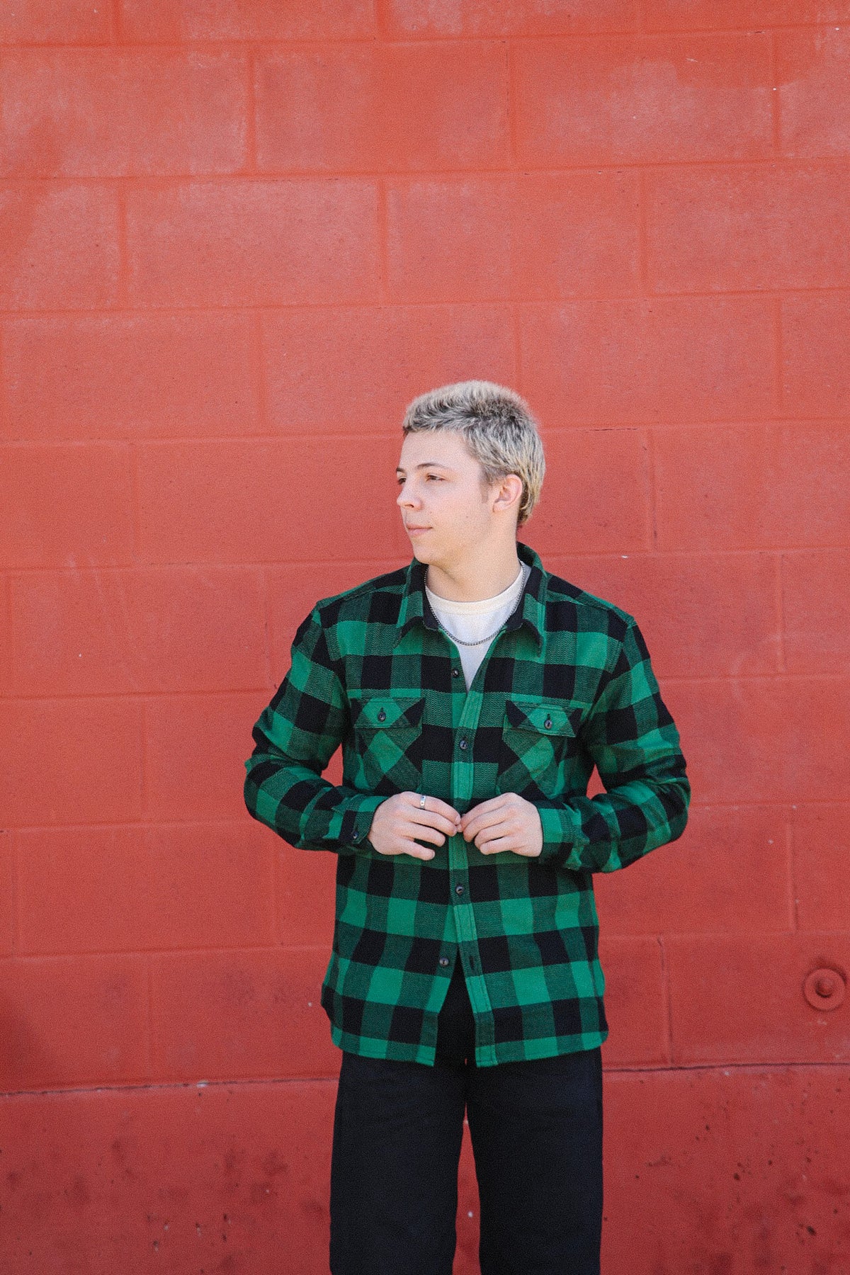 Young man wearing green and black plaid shirt standing against red brick wall