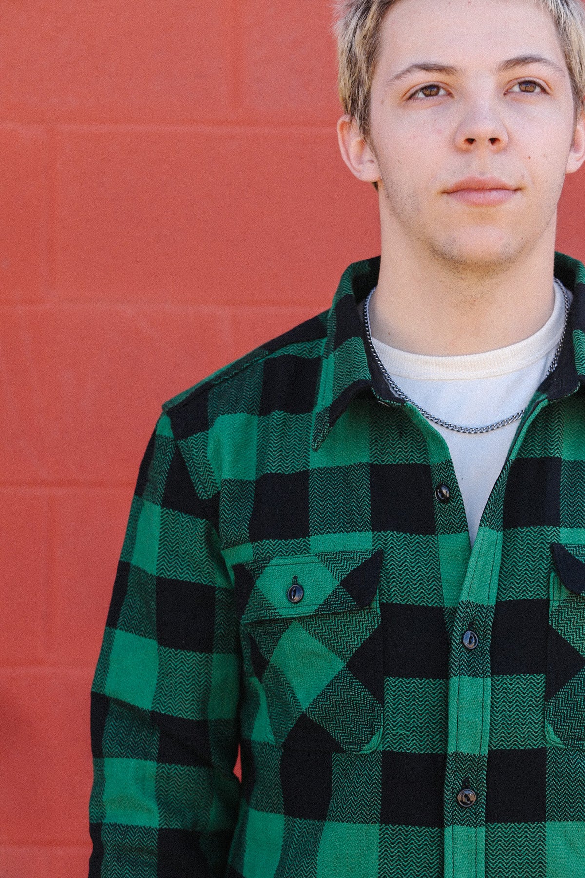 Young man in green and black checkered shirt with chain necklace against red brick wall