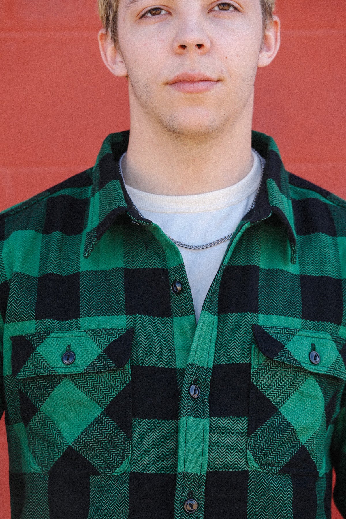 Close-up of a young man wearing a green and black plaid flannel shirt over a white t-shirt with a silver chain necklace against a red background
