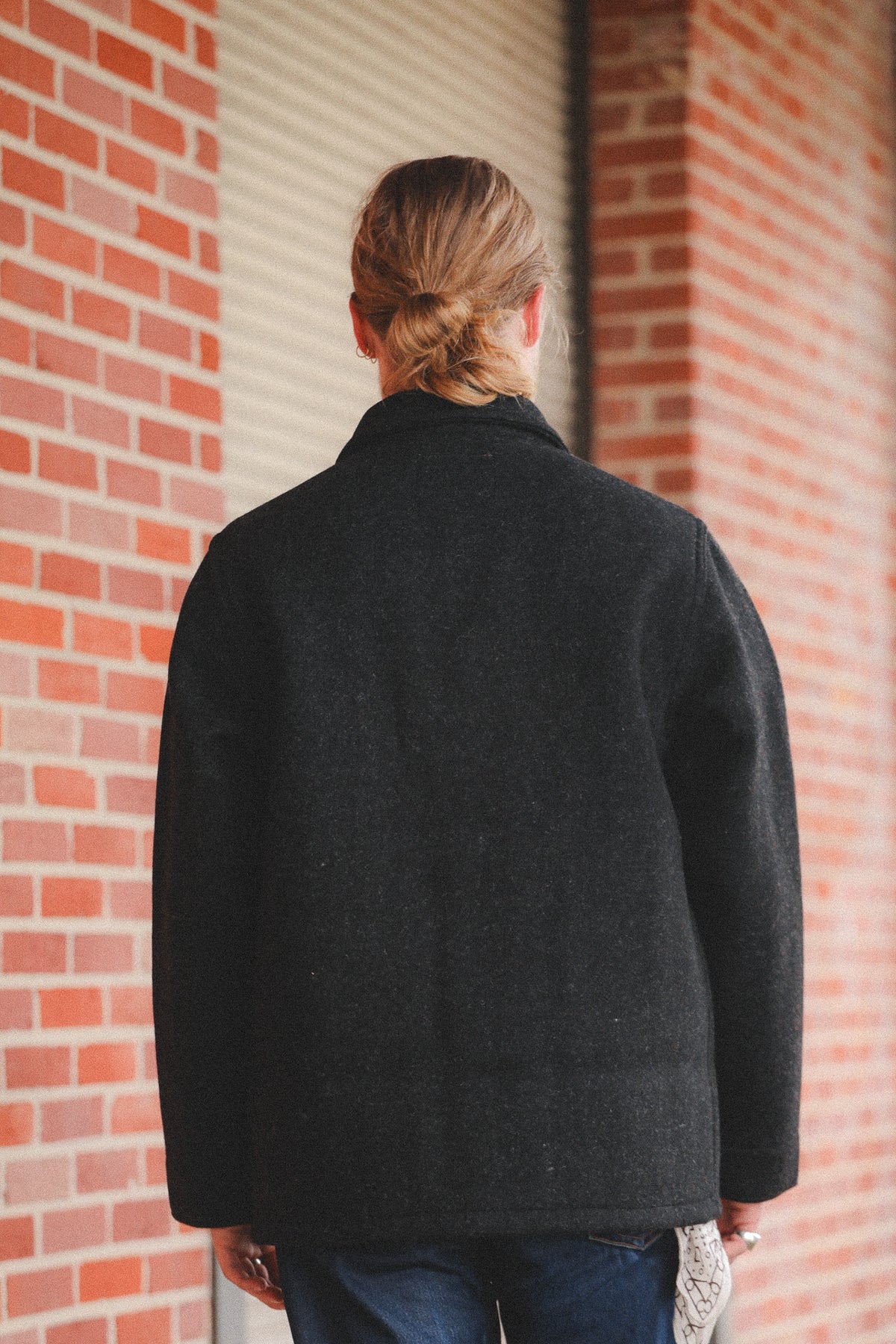 A person  wearing a dark charcoal gray wool-blend coat, standing in front of a red brick wall and a metal roll-up door.