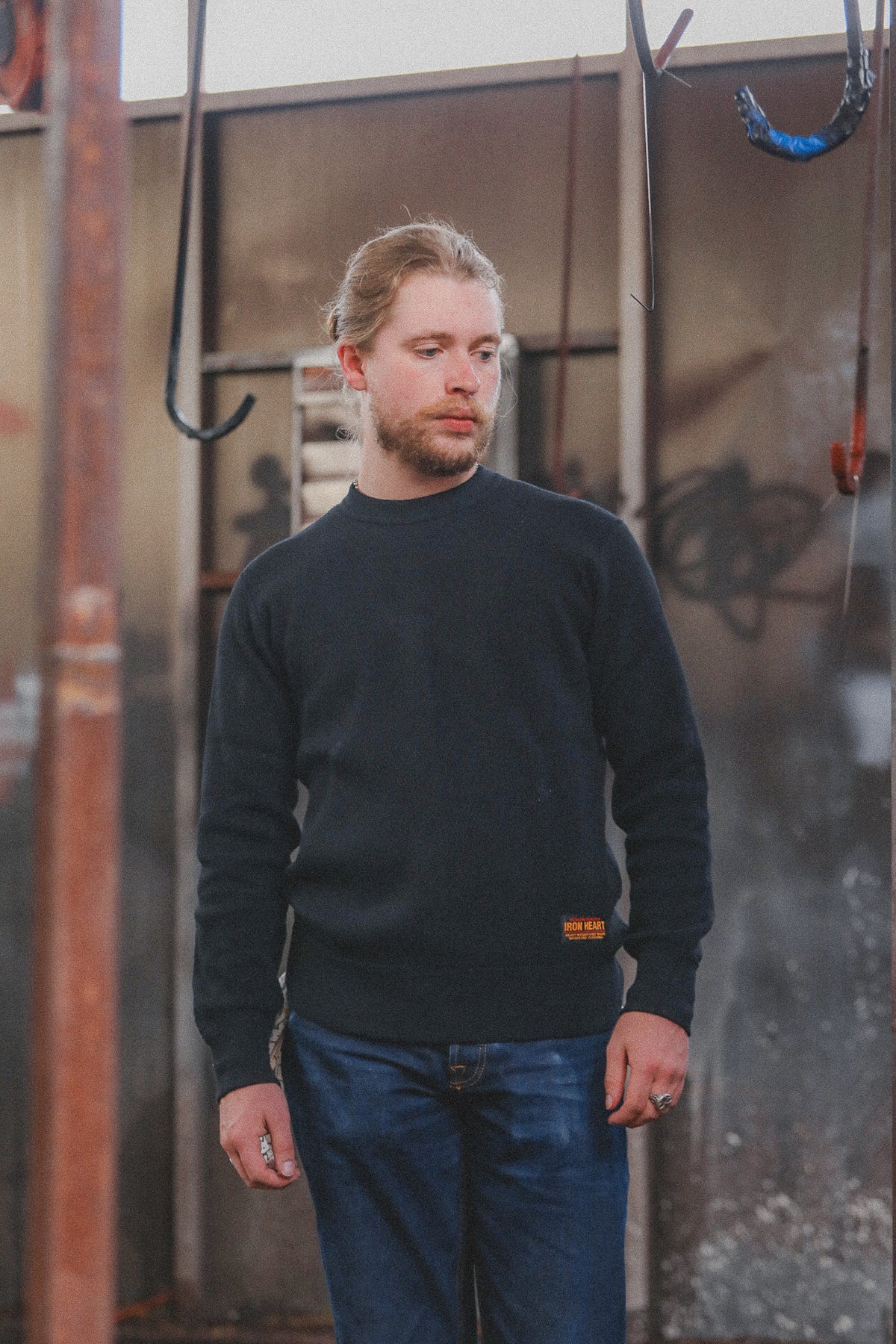 A man wearing a dark navy blue crewneck sweater and blue jeans, standing in front of a rustic, industrial metal wall with hanging hooks.