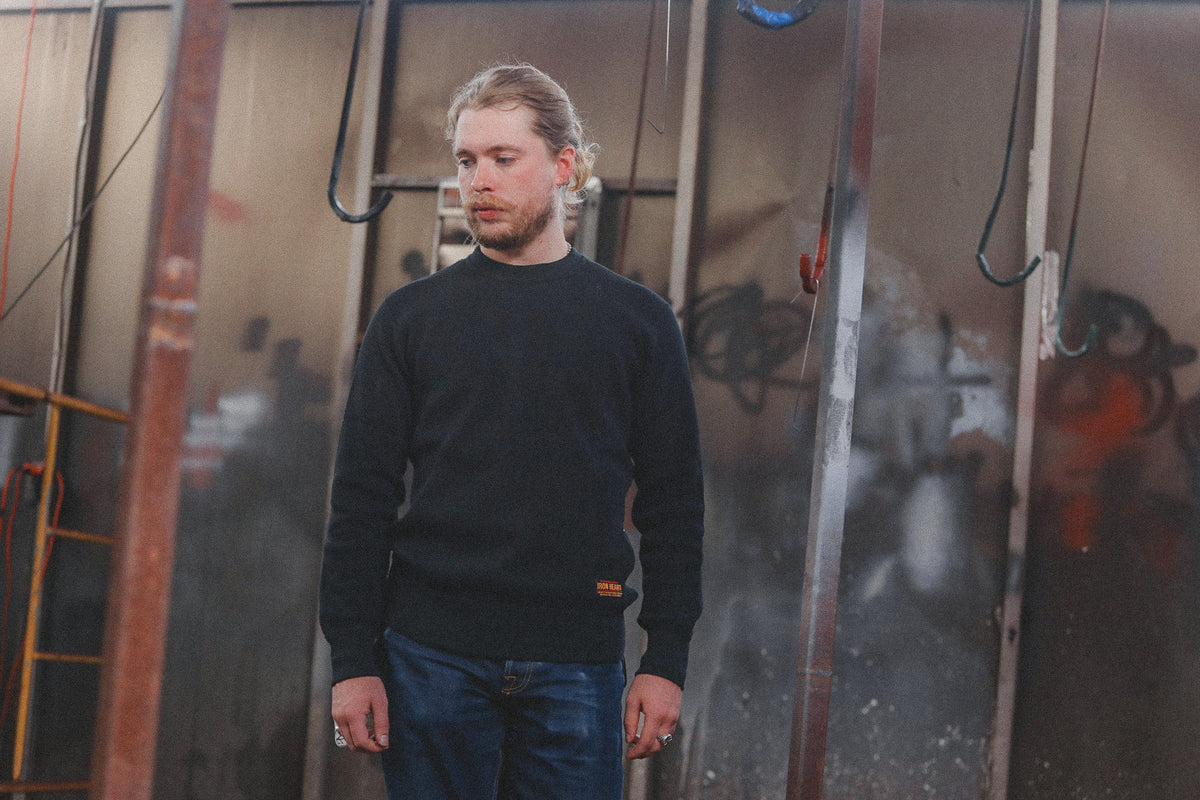 A man with blonde hair tied back stands wearing a black crewneck sweater and blue jeans in front of a graffiti-covered metal wall.