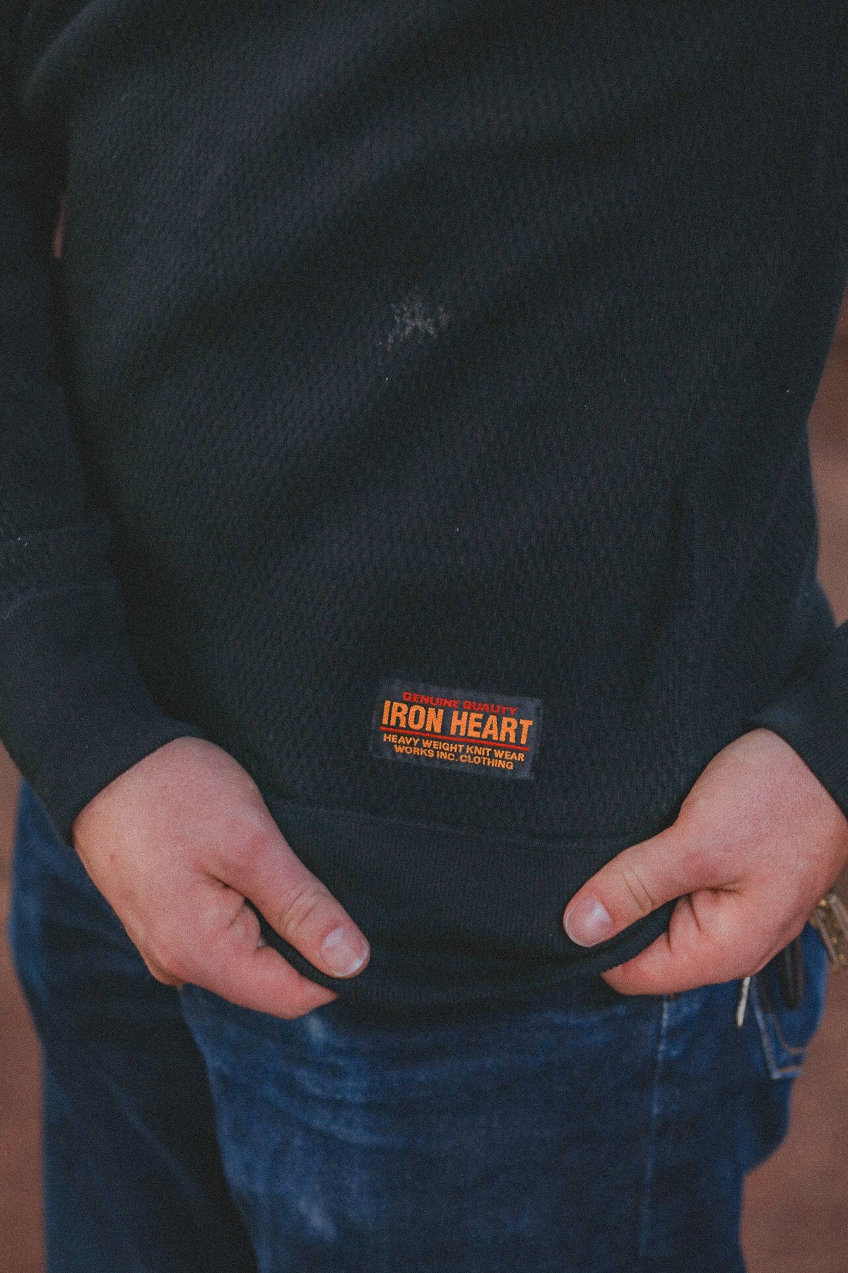 A close-up of a person wearing a black ribbed knit sweater with a small orange and white 'IRON HEART' brand patch on the front, paired with blue jeans.