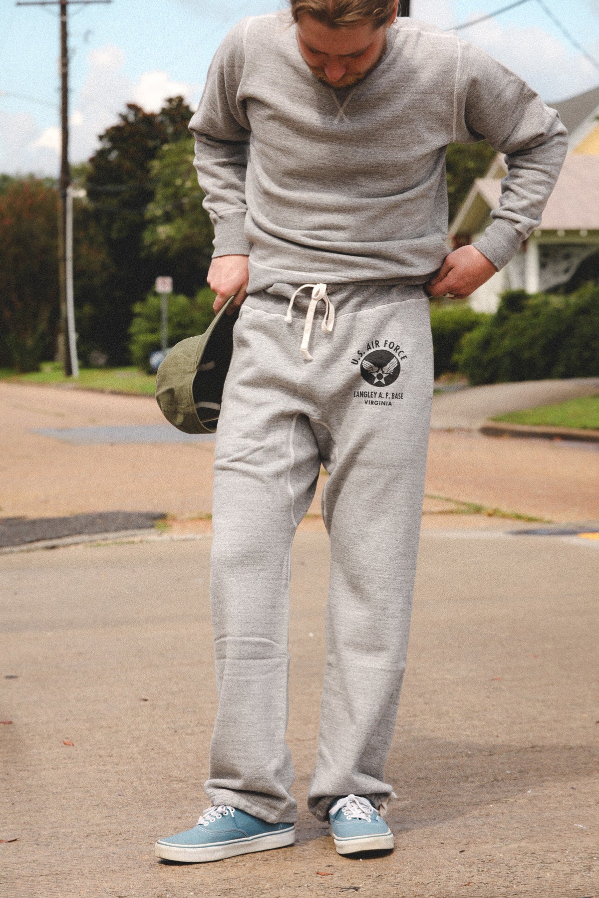 A man wearing a heather gray sweatshirt and matching gray sweatpants with a black graphic print on the thigh, standing on an outdoor street holding a green cap.